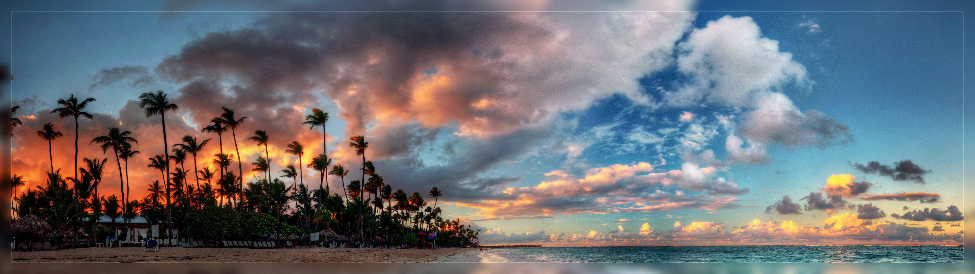 3840x1080 HD dual monitor 桌布 壁紙 — 充滿活力的日落將天空染成火熱的橙色和粉紅色，映照在棕櫚樹剪影排列的熱帶海灘上。