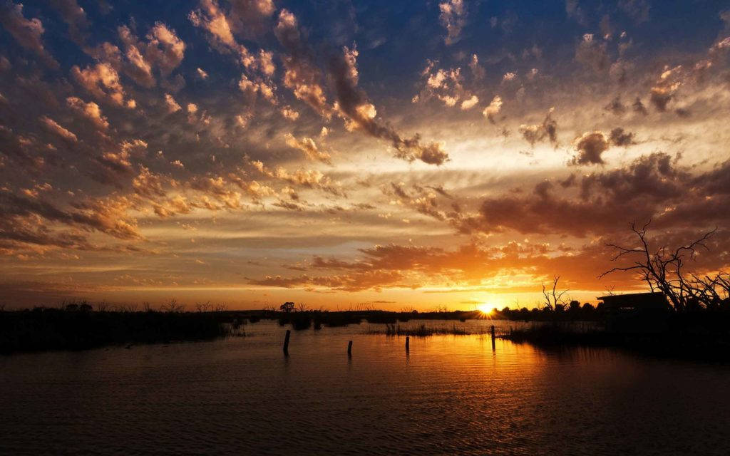 Sunrise Nature 桌布 壁紙 — 充滿活力的日落將天空染成火熱的橙色和深藍色，雲彩倒映在平靜的水面上，樹木和柱子的剪影清晰可見。