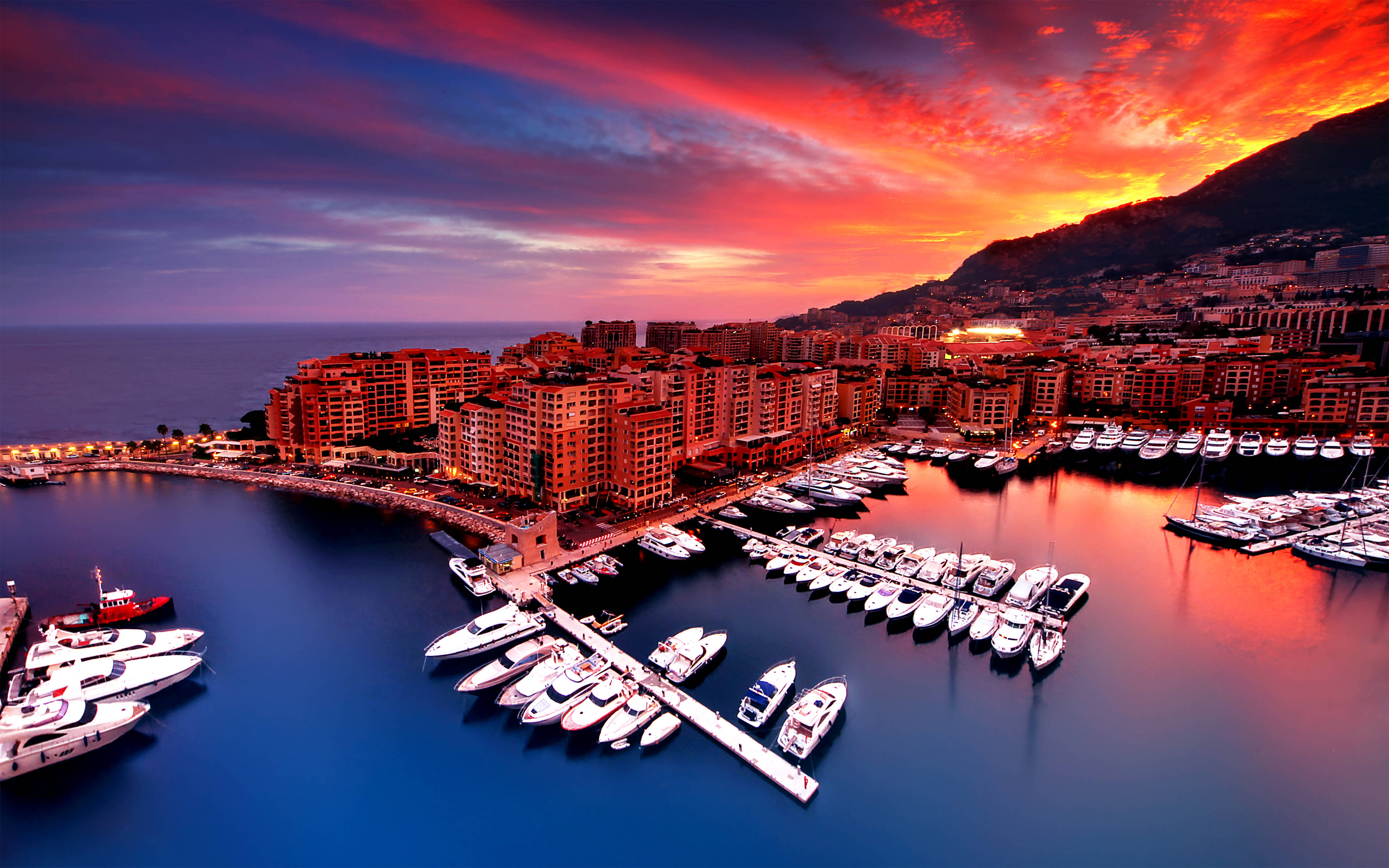 Monaco 桌布 壁紙 — 充滿活力的日落將天空染成火紅和橙色，映照著一個停滿豪華遊艇和燈火通明建築物的沿海港口。