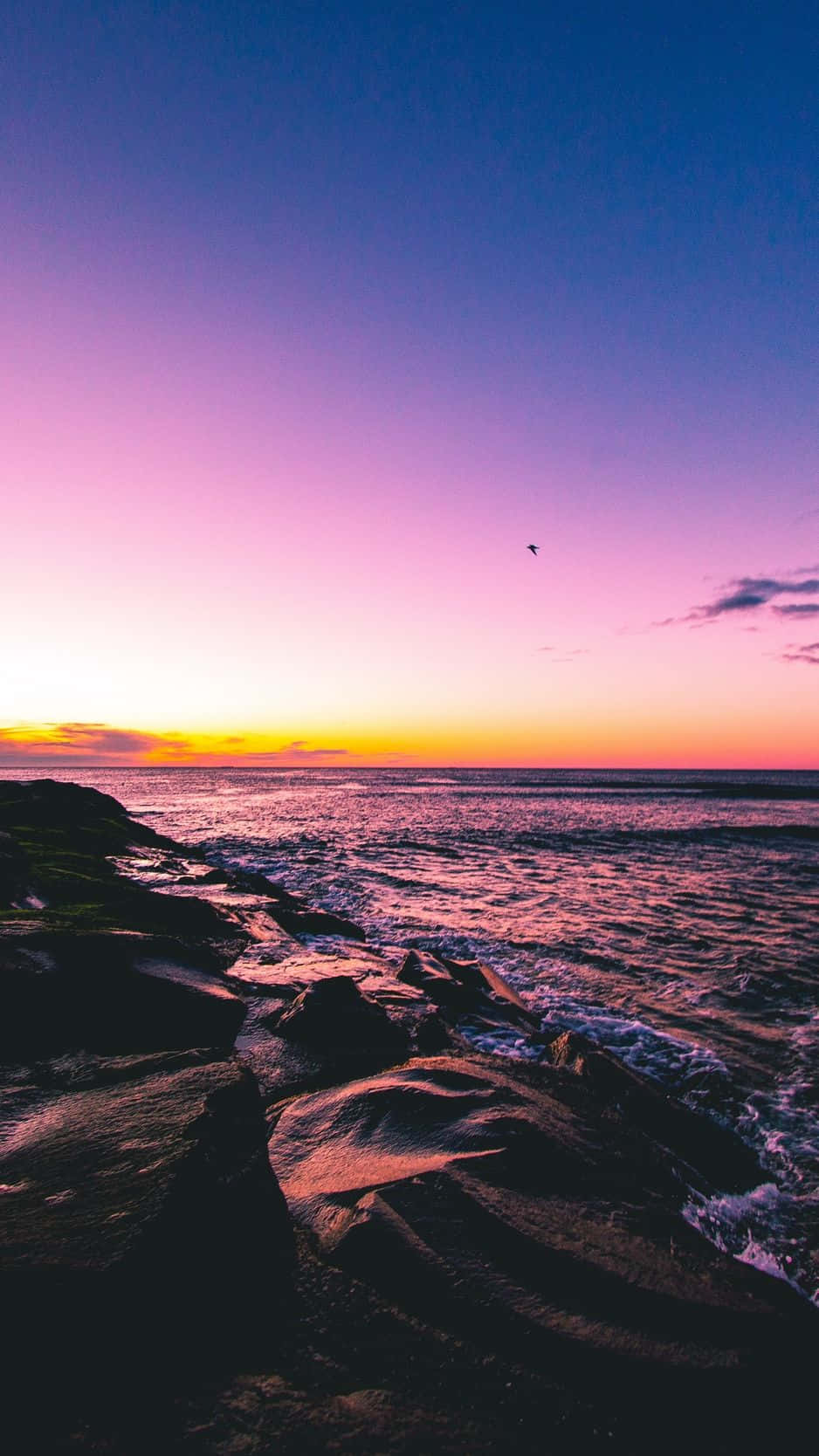 Sunset Beach iPhone 桌布 壁紙 — 充滿活力的日落將天空染成粉紅色、紫色和橙色，海浪拍打著黑暗的岩石海岸。