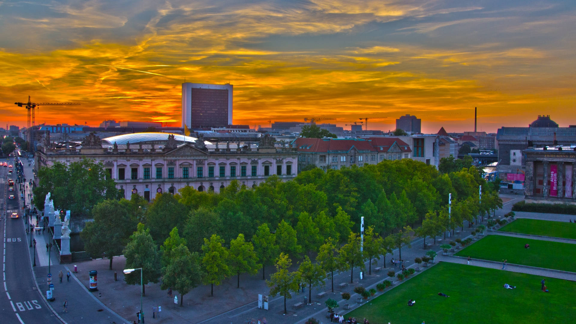 Berlin 桌布 壁紙 — 充滿活力的日落將歐洲城市景觀沐浴在金色光芒中，樹木和歷史建築環繞著繁華的城市公園。