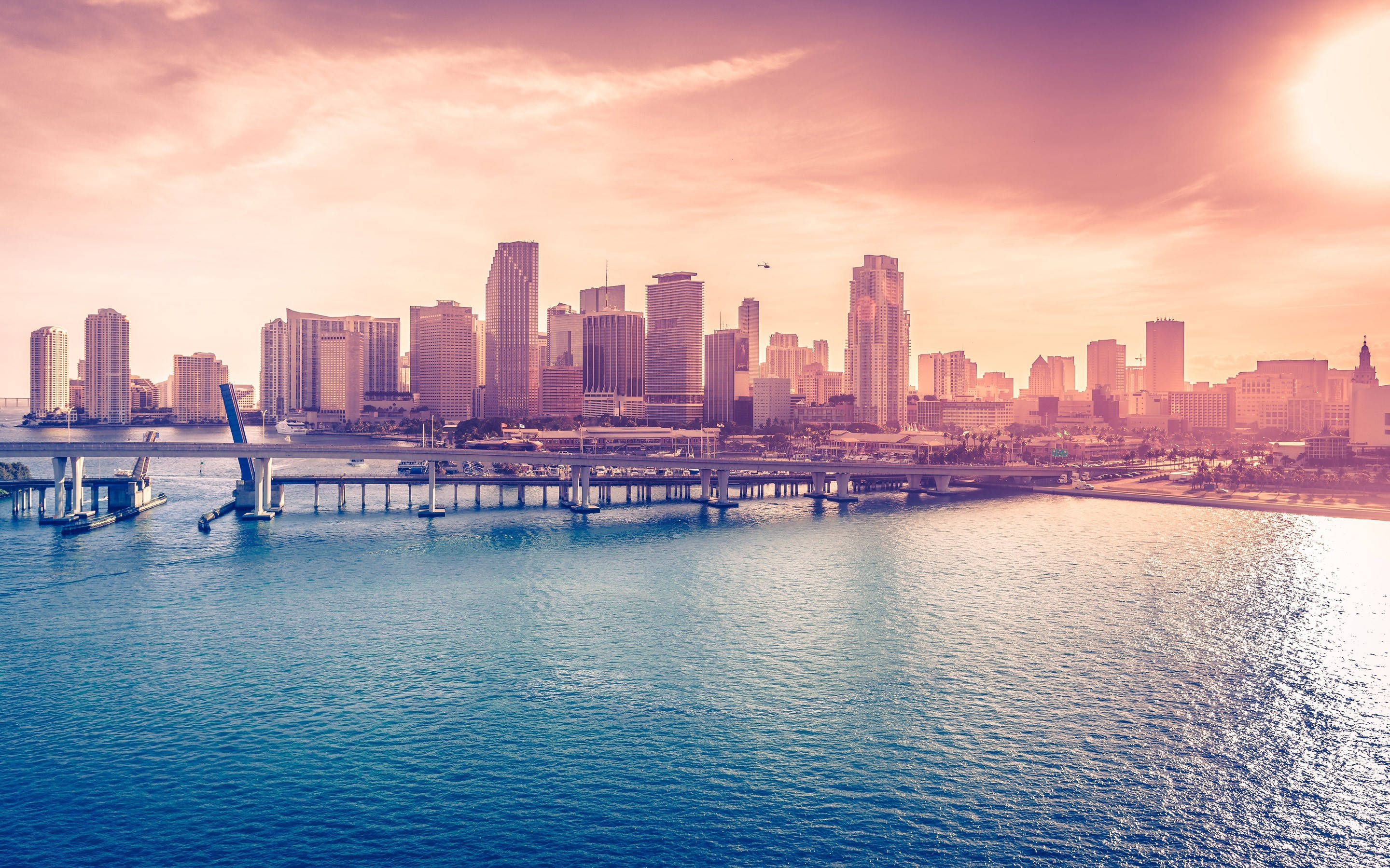 Miami 桌布 壁紙 — 充滿活力的日落將現代城市天際線和海濱大橋沐浴在溫暖的粉紅色和橙色色調中，映照在平靜的藍色水面上。