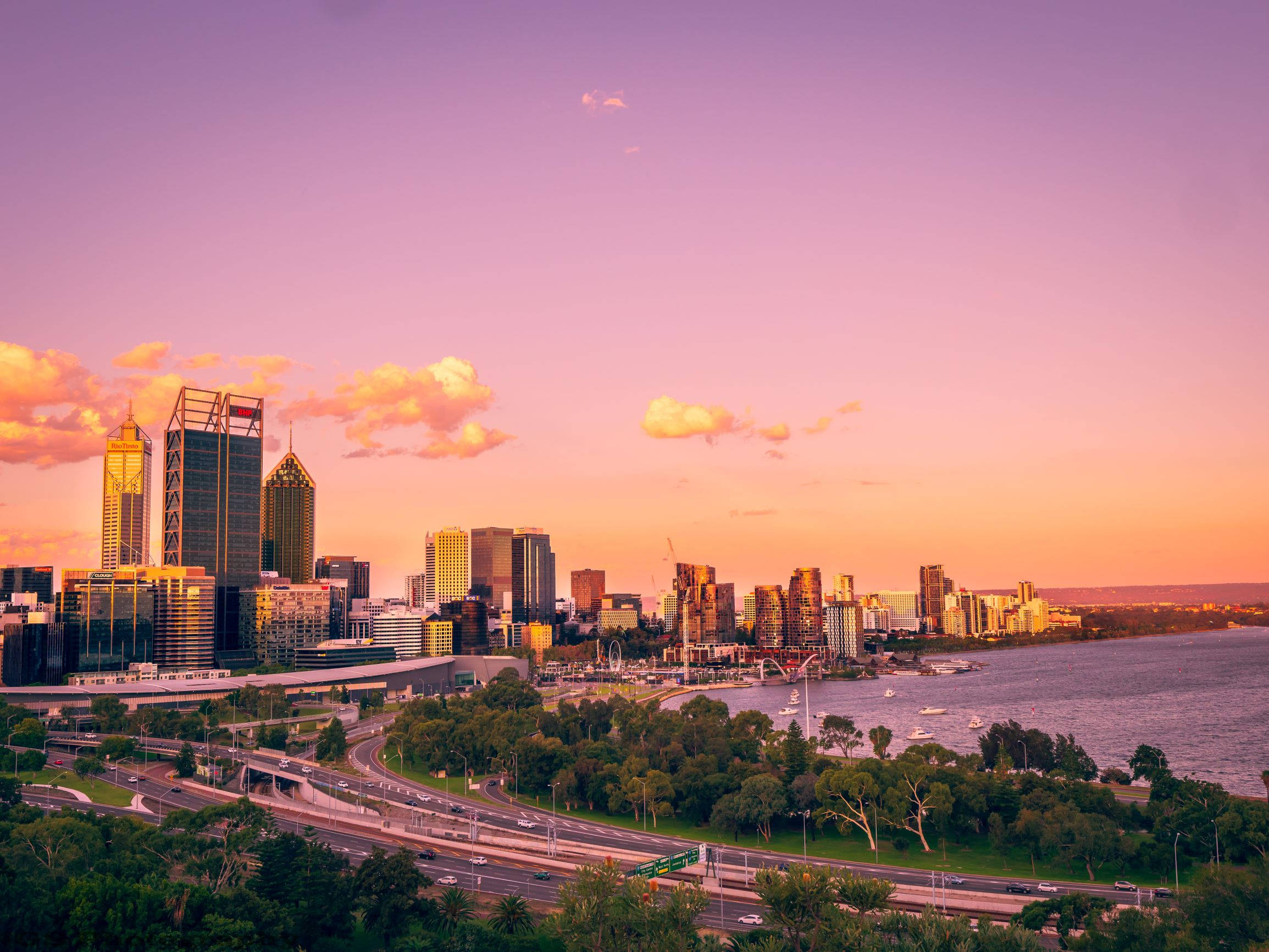 Perth 桌布 壁紙 — 充滿活力的日落將現代城市天際線和蜿蜒的高速公路沐浴在平靜的河流旁，呈現溫暖的橙色和紫色色調。