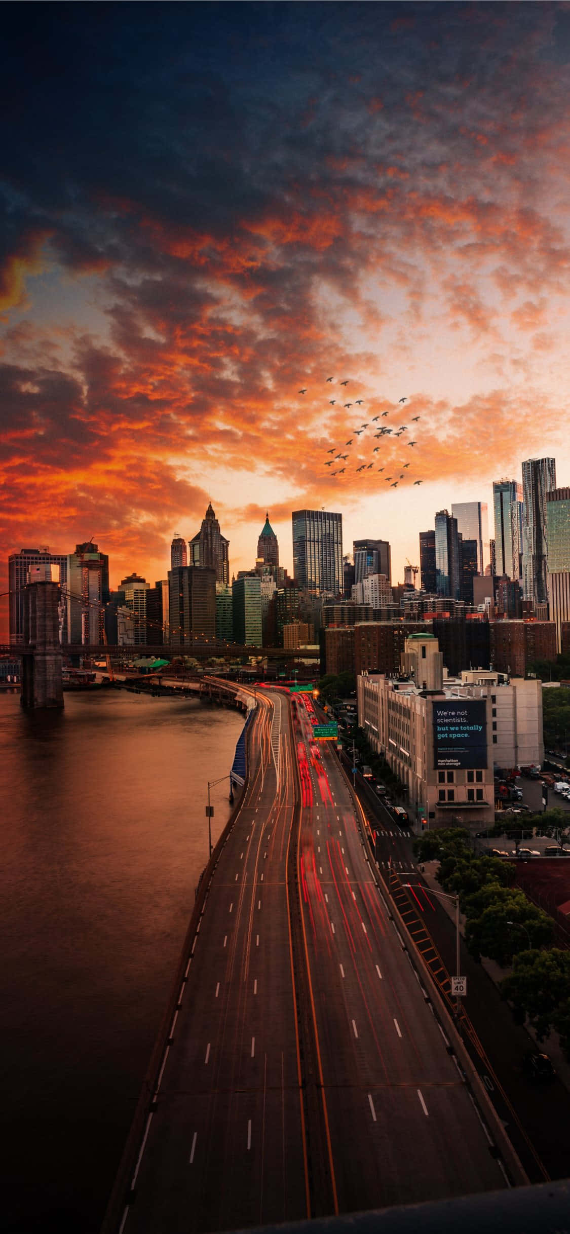 New York 桌布 壁紙 — 充滿活力的日落將紐約天際線染成火熱的橙色和紅色，河邊高速公路上的車流燈光形成一道道流光。