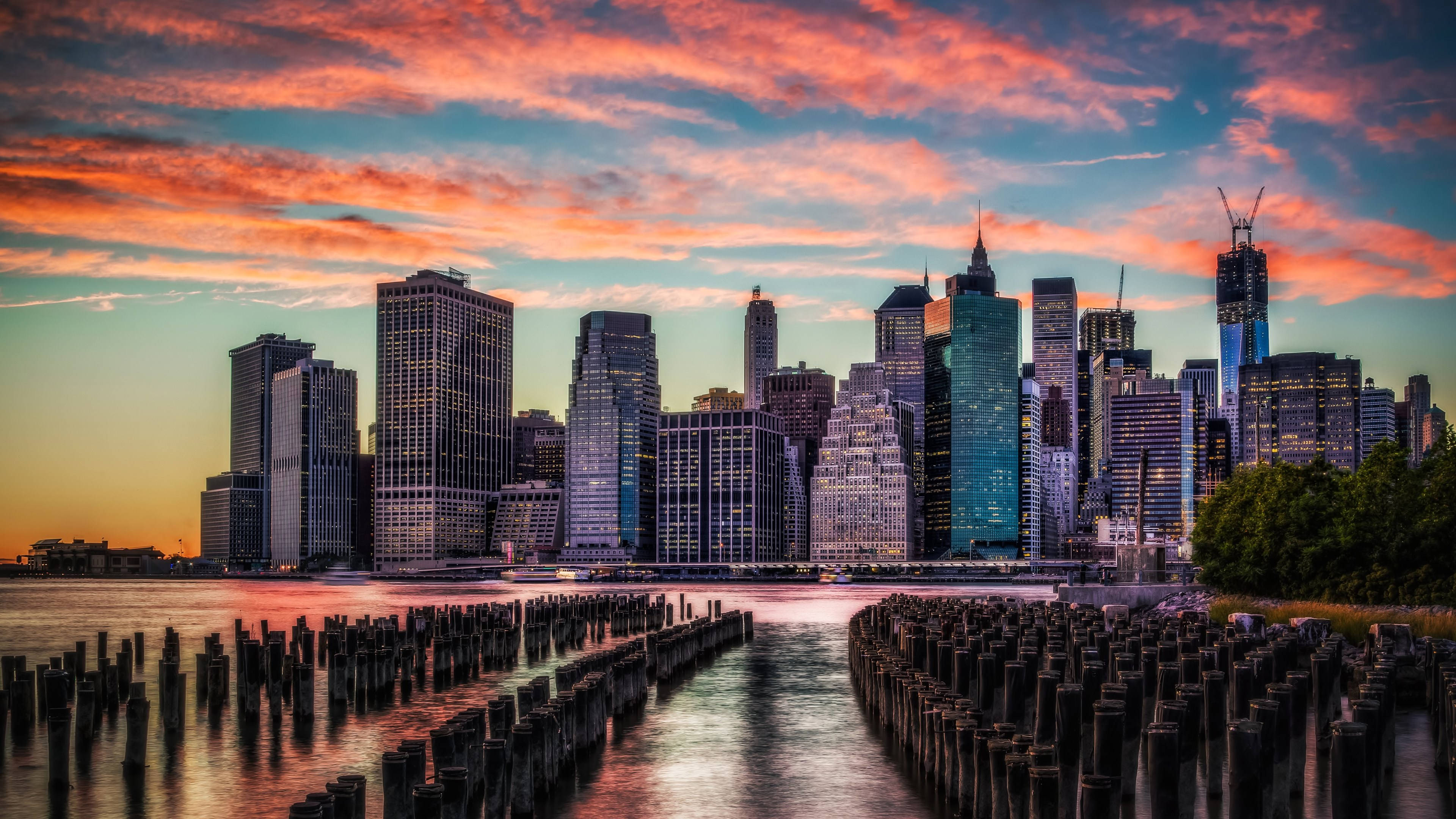 New York 桌布 壁紙 — 充滿活力的日落將紐約天際線染成火熱的橙色和粉紅色，倒映在水面上，前景有風化的木樁。