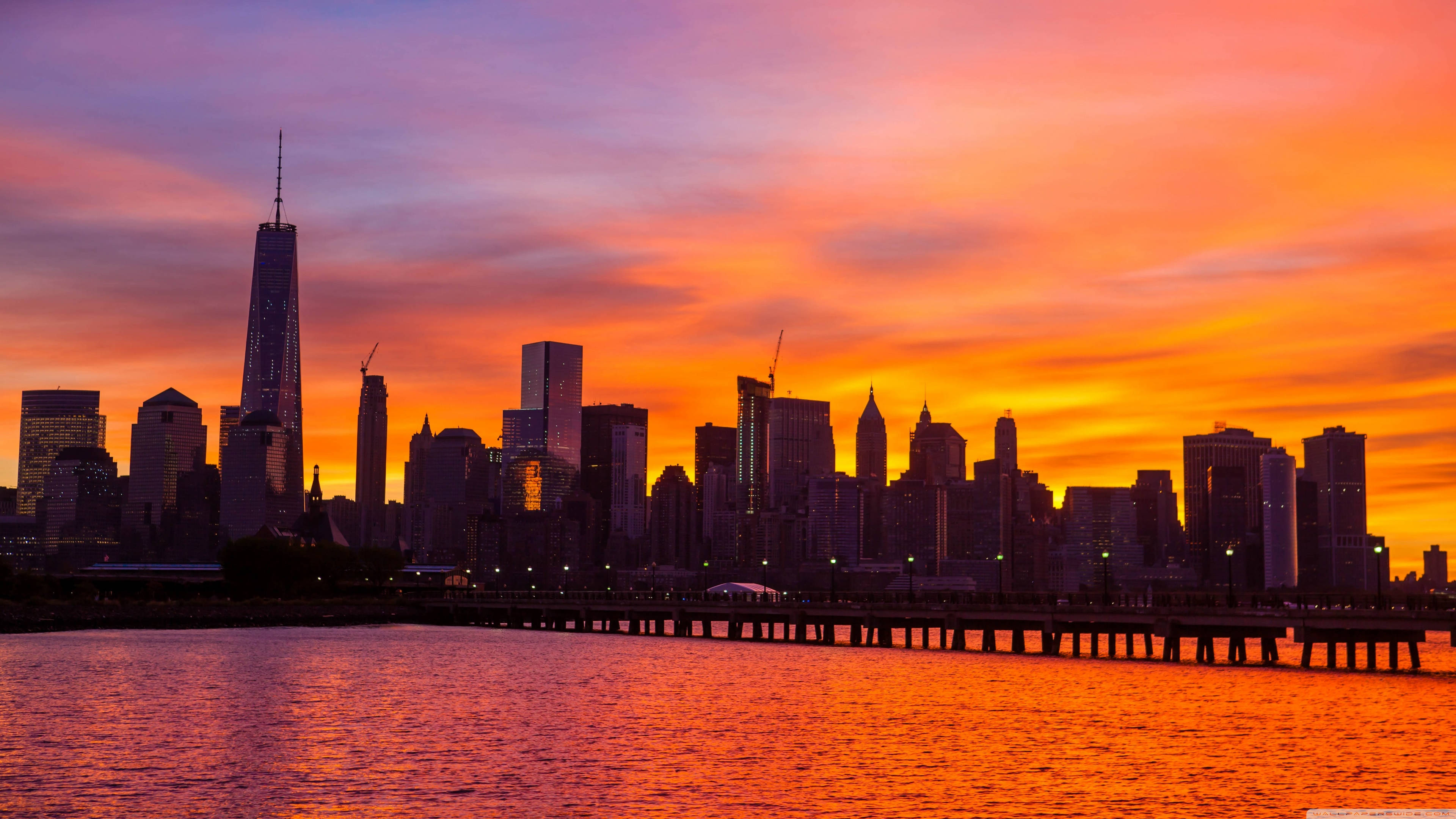 4K New York City 桌布 壁紙 — 充滿活力的日落將紐約市的天際線染成火熱的橙色和粉紅色調，倒映在下方水面上。