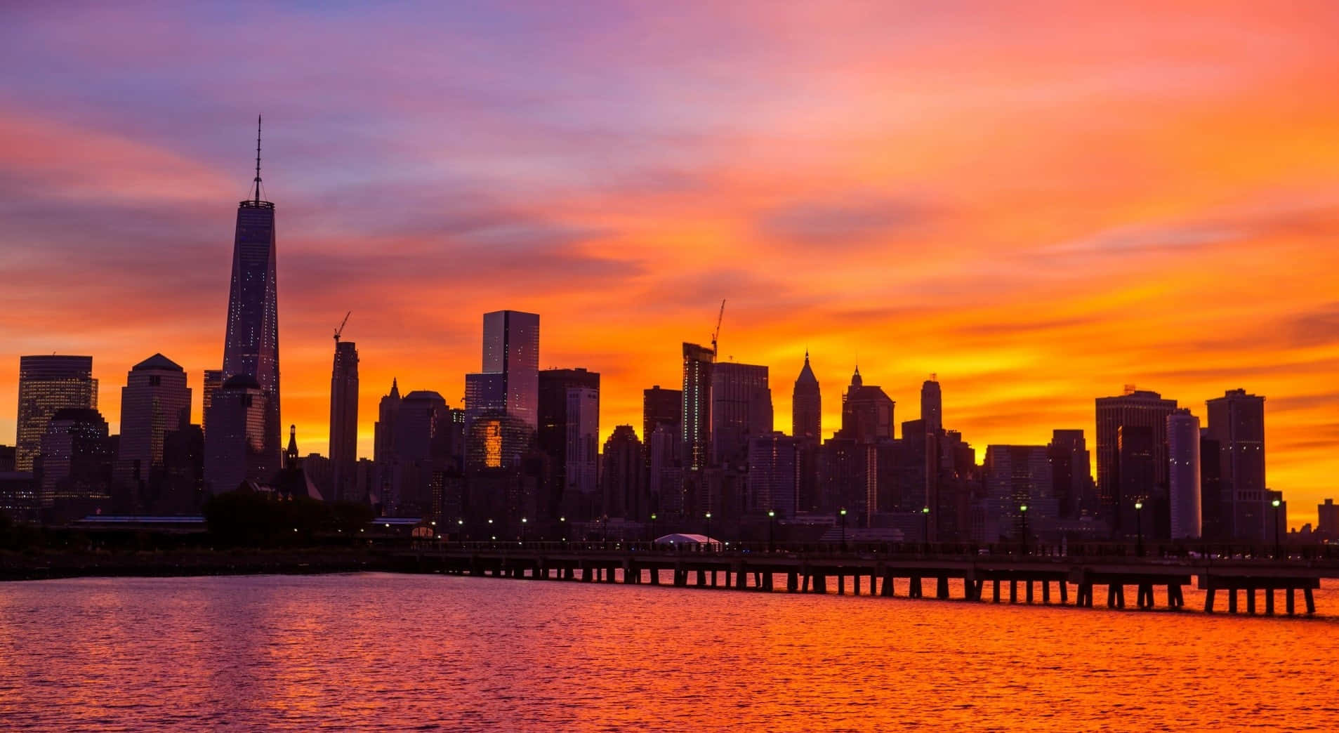 Nyc 桌布 壁紙 — 充滿活力的日落將紐約市的天際線染成火熱的橙色和紫色色調，倒映在下面的水面上。