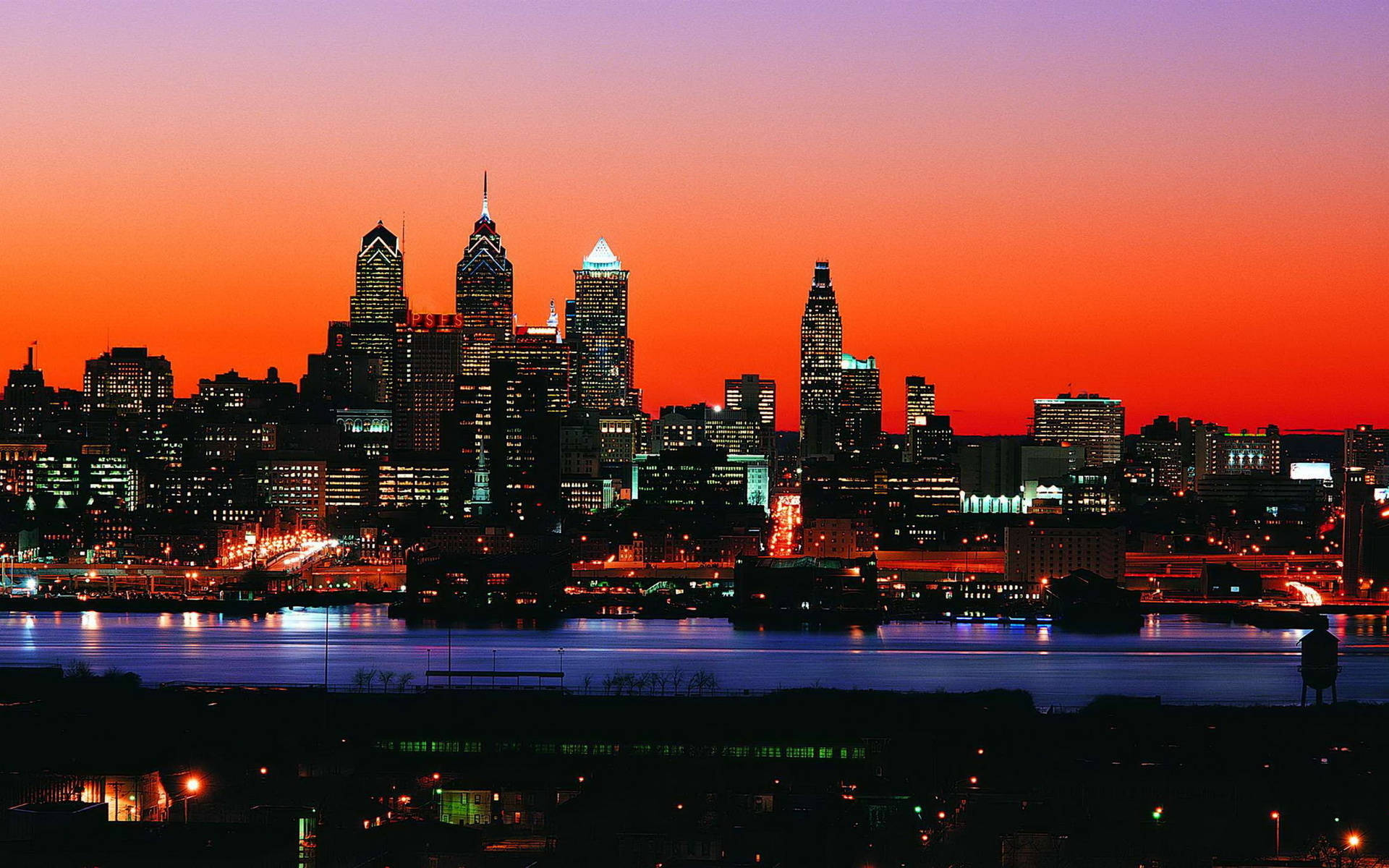 Philadelphia 桌布 壁紙 — 充滿活力的日落將費城天際線染成火熱的橙色和紫色，城市燈光倒映在下面的河流中。
