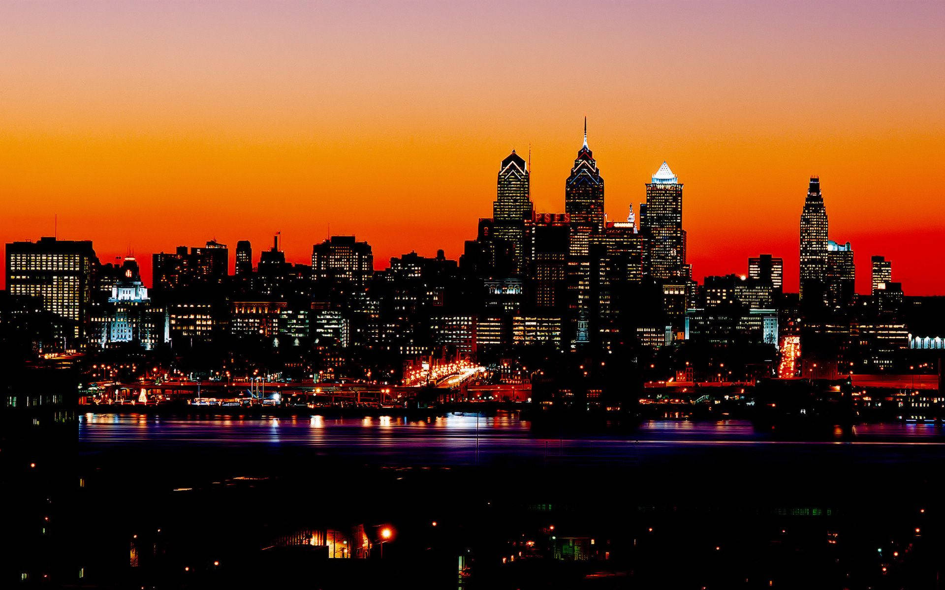 Philadelphia 桌布 壁紙 — 充滿活力的日落將費城天際線染成火熱的橙色和粉紅色，城市的燈光倒映在下方黑暗的河流中。