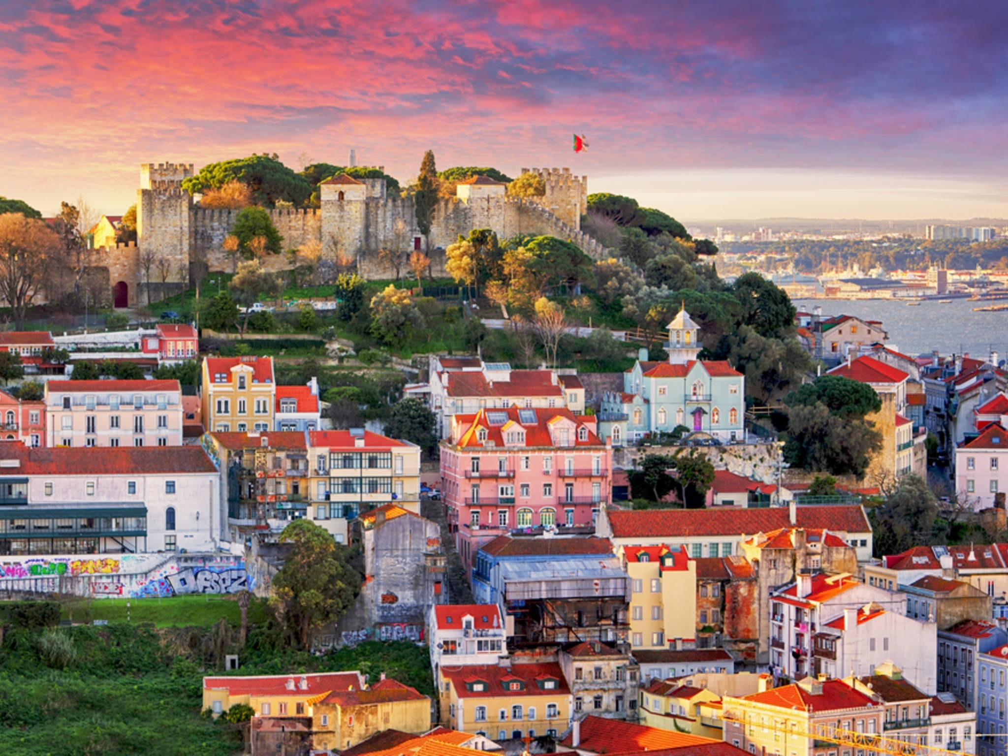 Portugal 桌布 壁紙 — 充滿活力的日落將里斯本的歷史城堡和色彩繽紛的瓷磚屋頂沐浴在溫暖的粉紅色和橙色色調中。