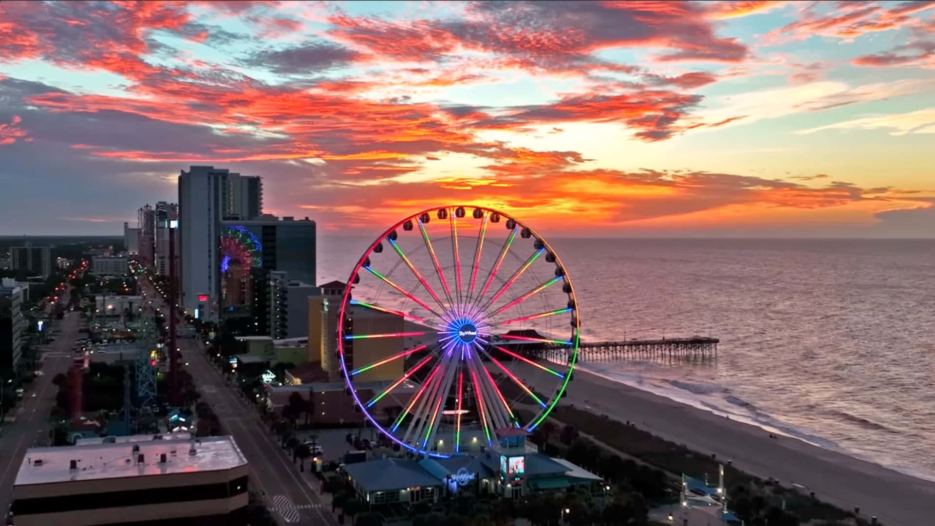 Myrtle Beach 桌布 壁紙 — 充滿活力的日落用火熱的色調描繪天空，在海灘城市景觀旁邊的發光摩天輪上方。