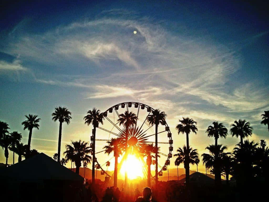Coachella 桌布 壁紙 — 充滿活力的日落襯托出巨型摩天輪和棕櫚樹的剪影，天空戲劇性變幻，帶有縷縷雲彩和一輪可見的月亮。