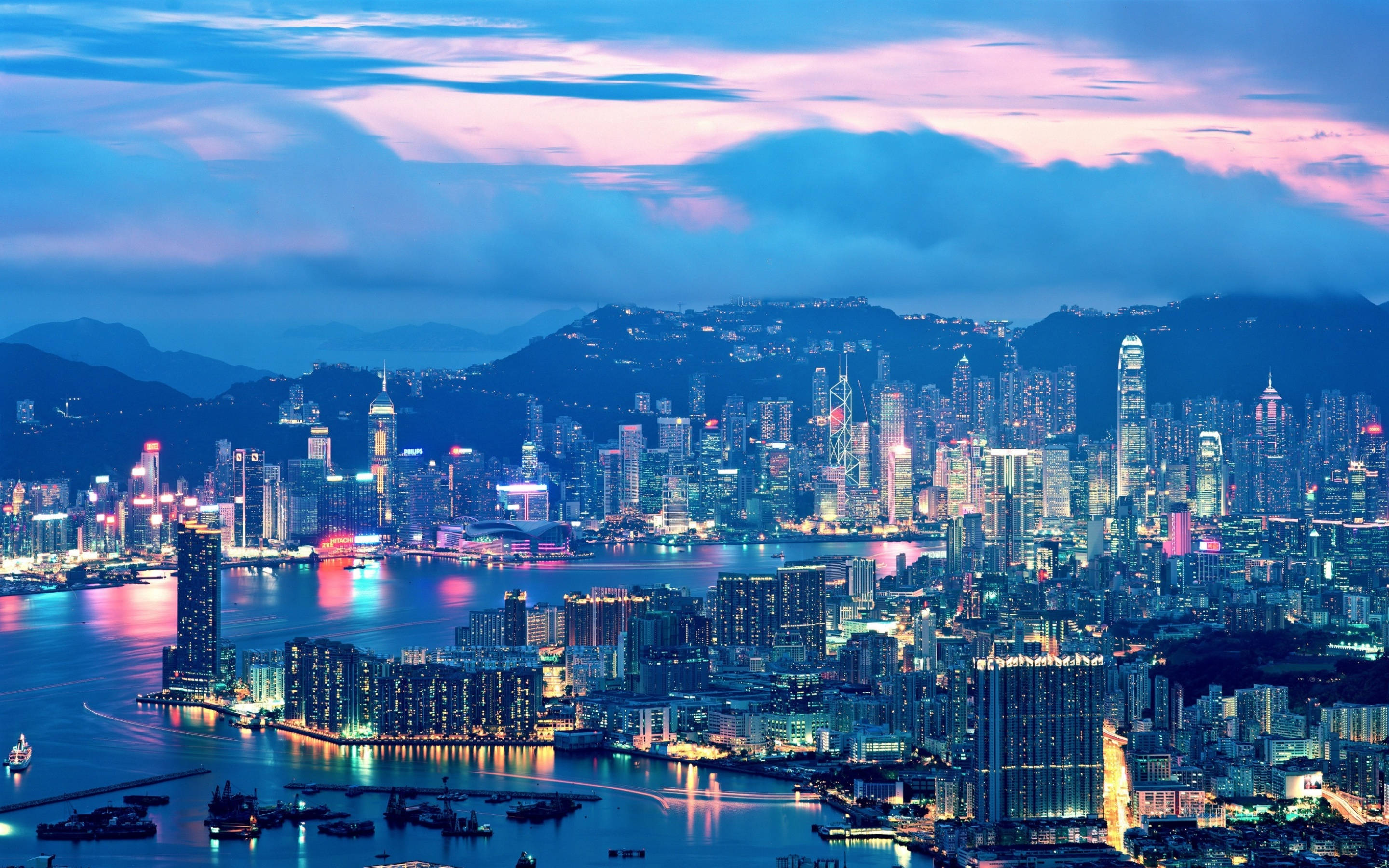 lights 桌布 壁紙 — 充滿活力的暮色城市景觀，發光的摩天大樓倒映在水面上，周圍環繞著山脈，天空呈現粉紅色和藍色。