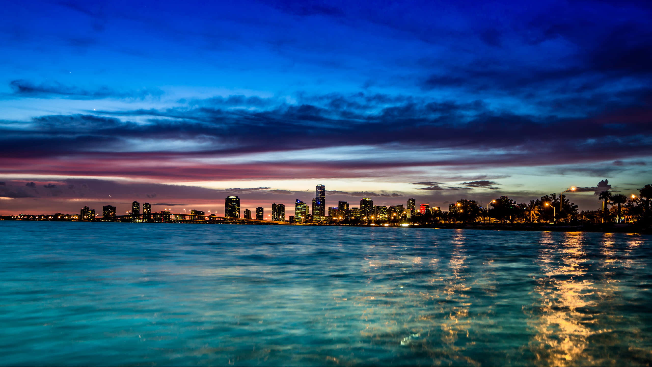 Miami 桌布 壁紙 — 充滿活力的暮色城市景觀，發光的摩天大樓倒映在平靜的藍綠色水面上，天空呈現戲劇性的紫色和藍色。