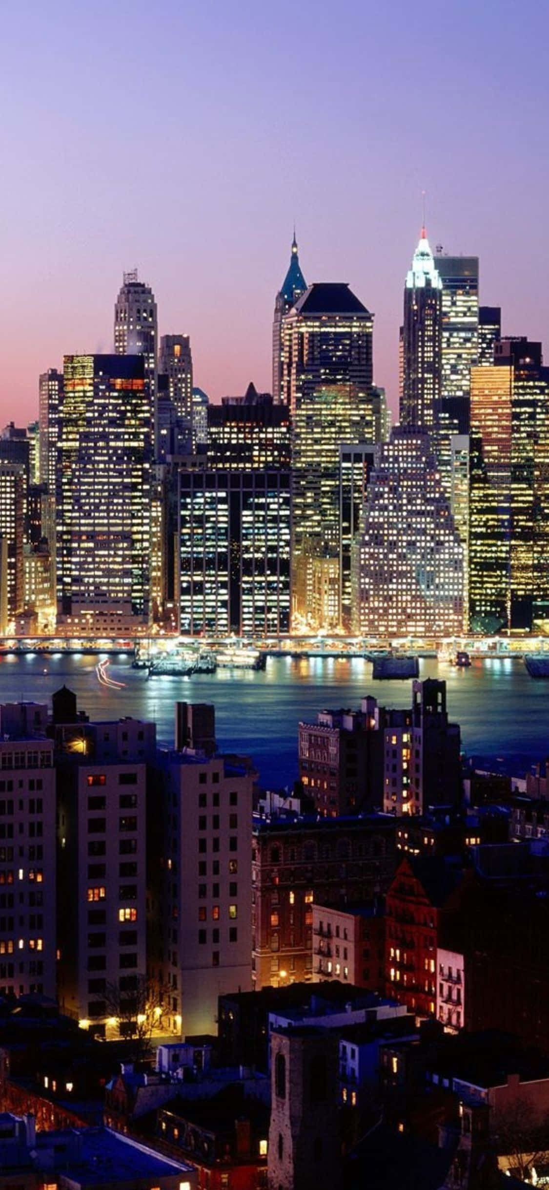 iPhone Xs Ny City 桌布 壁紙 — 充滿活力的暮色城市景觀，高聳的摩天大樓在紫色天空下閃耀，倒映在平靜的水面上。