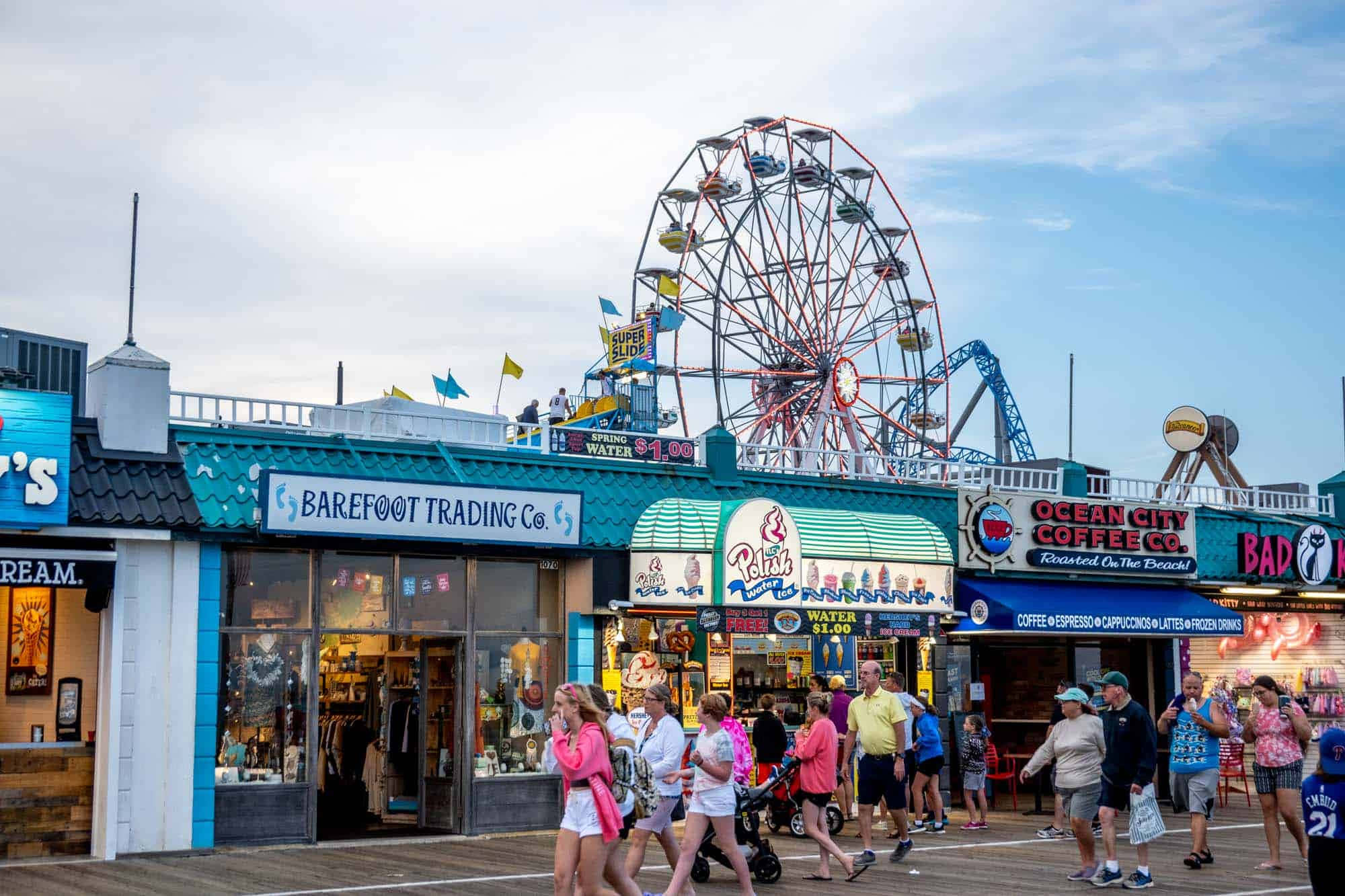 Boardwalk 桌布 壁紙 — 充滿活力的木板路場景，人們在色彩繽紛的商店和高聳的紅色摩天輪旁漫步，天空部分多雲。