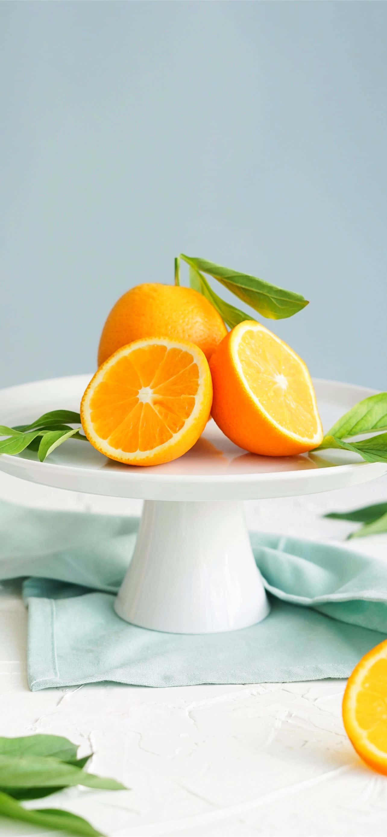 Lemon iPhone 桌布 壁紙 — 充滿活力的柳橙半邊和完整水果，帶有綠葉，放在淺藍色布上的白色蛋糕架上。