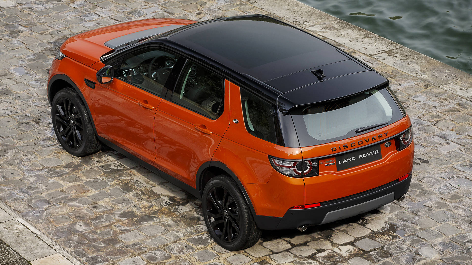 land rover iPhone 桌布 壁紙 — 充滿活力的橙色 Land Rover Discovery 停放在濕滑的鵝卵石上，旁邊是平靜的水面，柔和日光下。