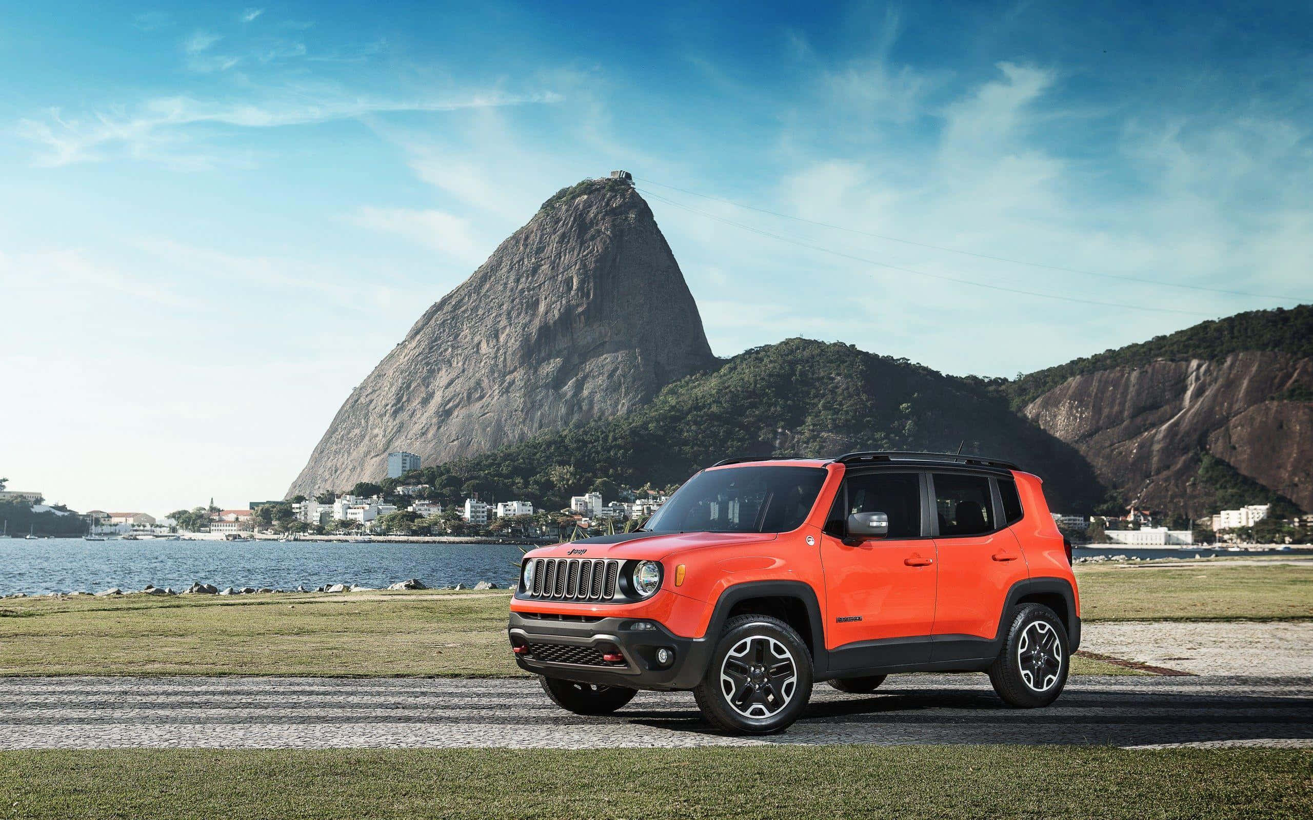 Jeep Renegade 桌布 壁紙 — 充滿活力的橙色Jeep Renegade停在草地近水處，背後是標誌性的糖麵包山在清澈藍天下聳立。