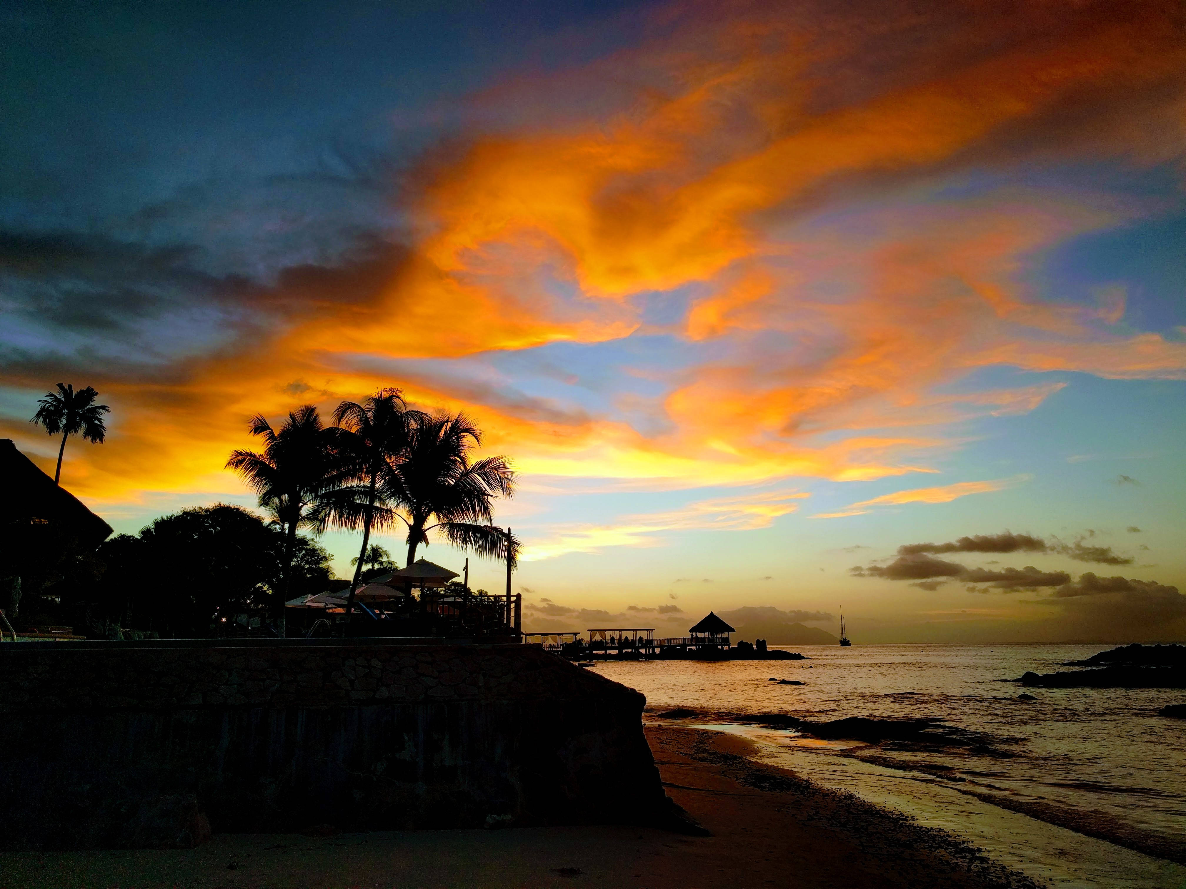 Seychelles 桌布 壁紙 — 充滿活力的橙色和藍色日落雲彩勾勒出棕櫚樹和碼頭在平靜海面上的剪影。