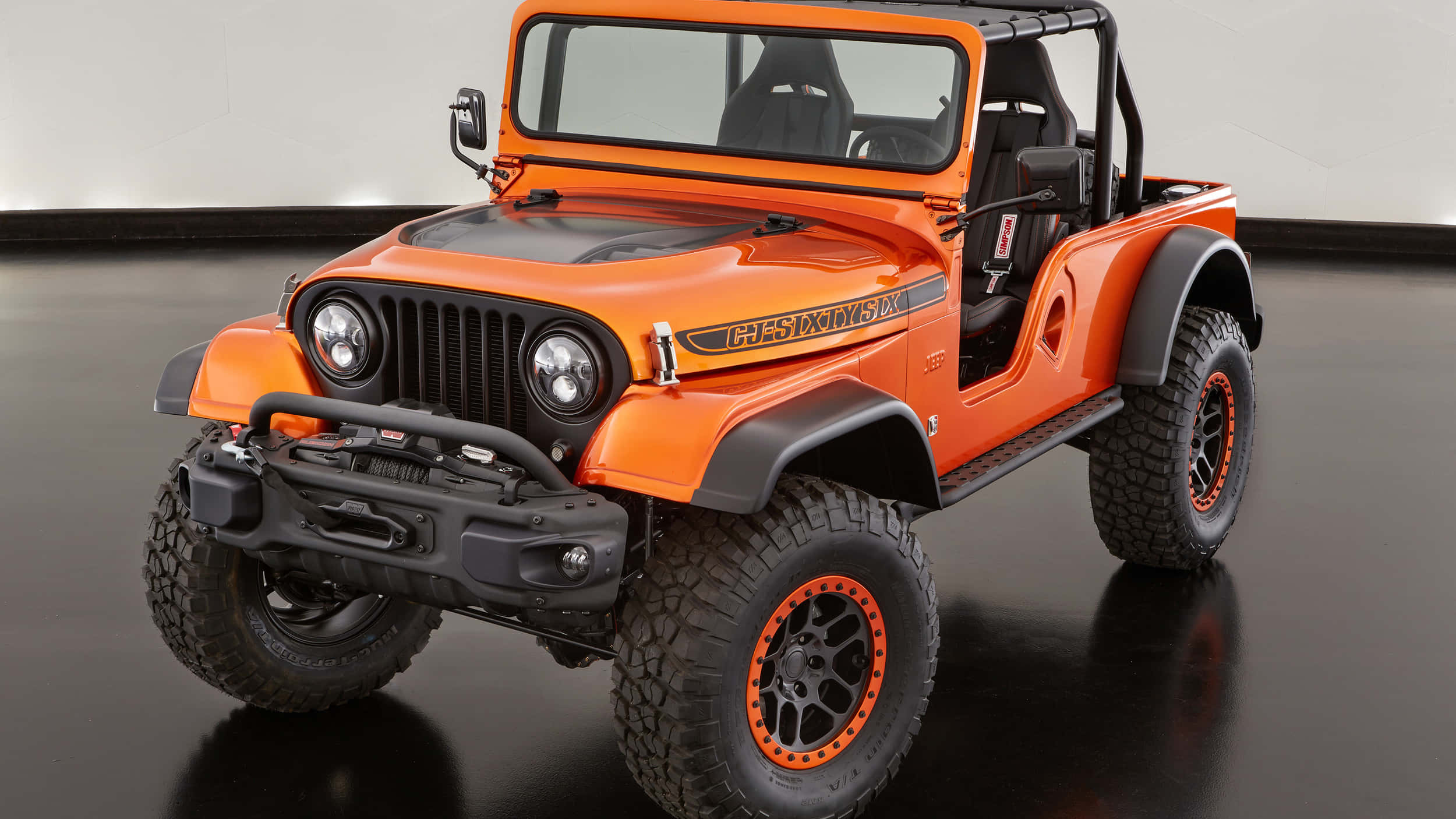 Jeep Cj 桌布 壁紙 — 充滿活力的橙色定制Jeep Wrangler，帶有黑色飾邊和大型越野輪胎，擺放在光亮的黑色工作室地板上。