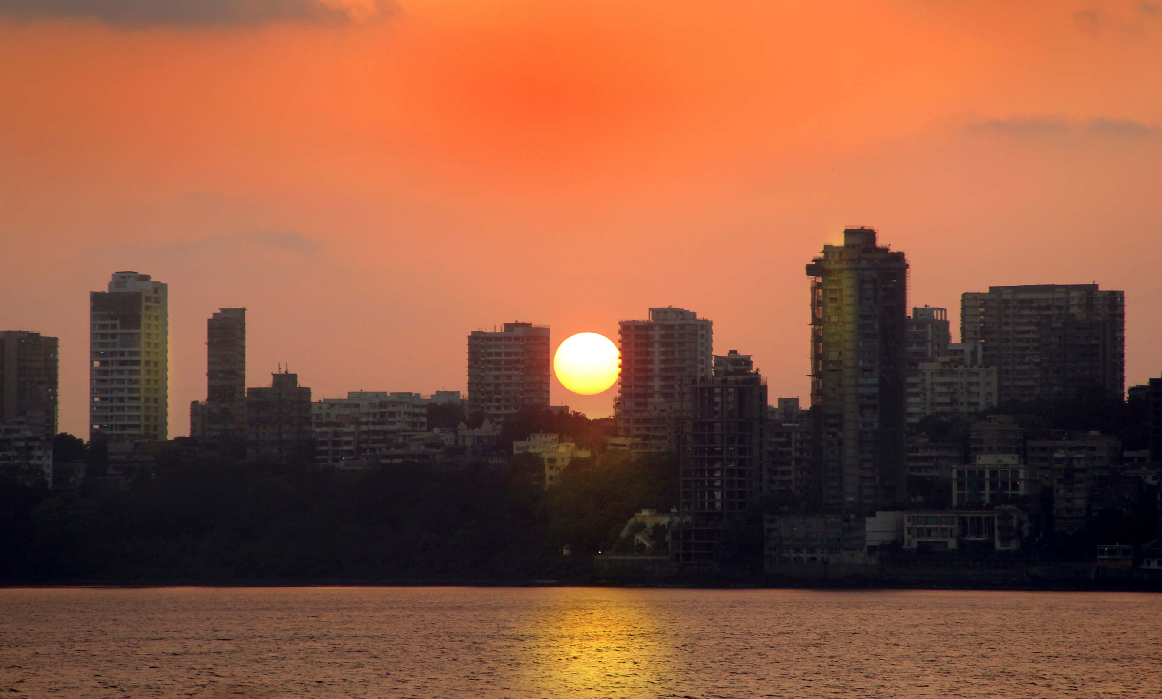 Mumbai 桌布 壁紙 — 充滿活力的橙色日落，在城市天際線後方閃耀，倒映在平靜的水面上。