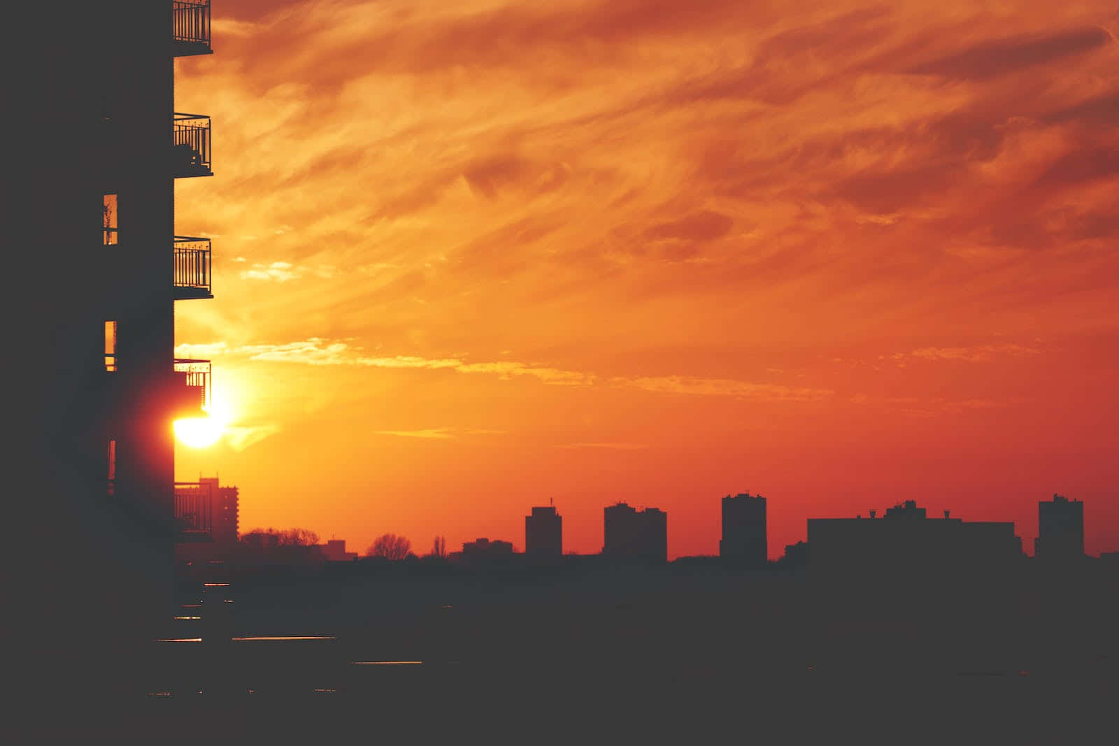 Cute Sunset 桌布 壁紙 — 充滿活力的橙色日落為城市天際線投下溫暖的光芒，左側有輪廓建築和一座突出的公寓大樓。