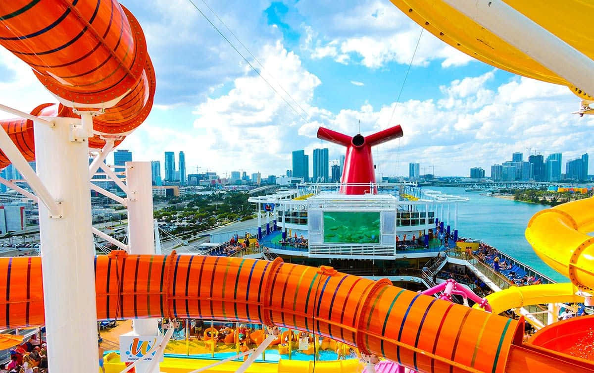 Carnival Vista 桌布 壁紙 — 充滿活力的橙色水上滑梯在繁忙的郵輪甲板上蜿蜒，紅色煙囪襯托著明亮的藍天和城市天際線。