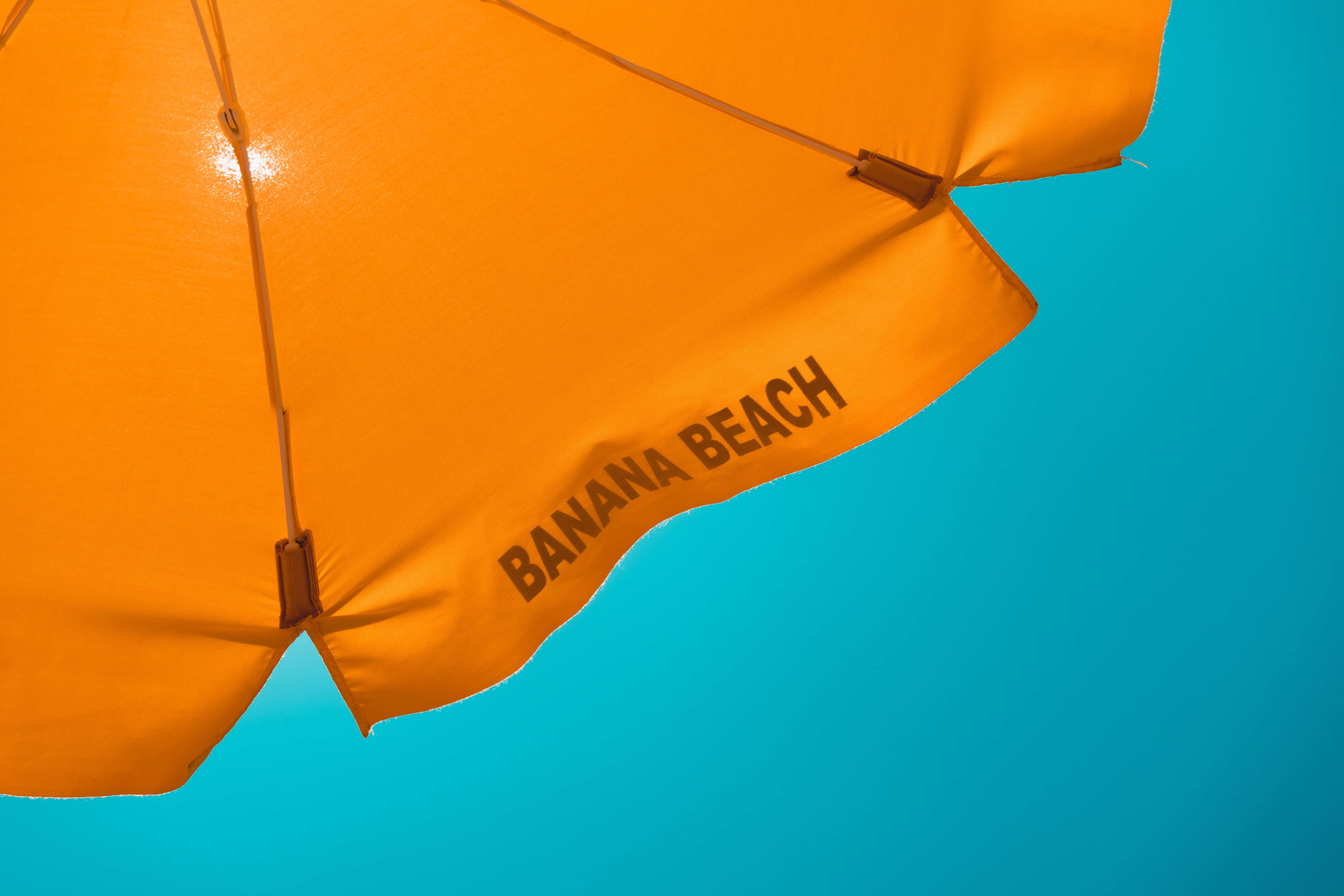 Beach Umbrella 桌布 壁紙 — 充滿活力的橙色沙灘傘，上面有「BANANA BEACH」字樣，背景是明亮的藍綠色天空。