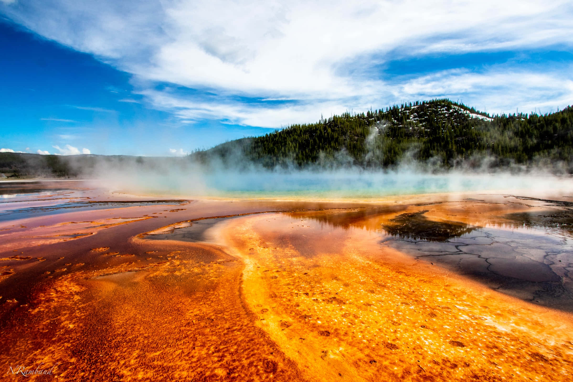 Yellowstone Geysers 桌布 壁紙 — 充滿活力的橙色礦物池在晴朗的藍天下，靠近樹木繁茂的山丘，冒著蒸汽，天空中有蓬鬆的雲朵。