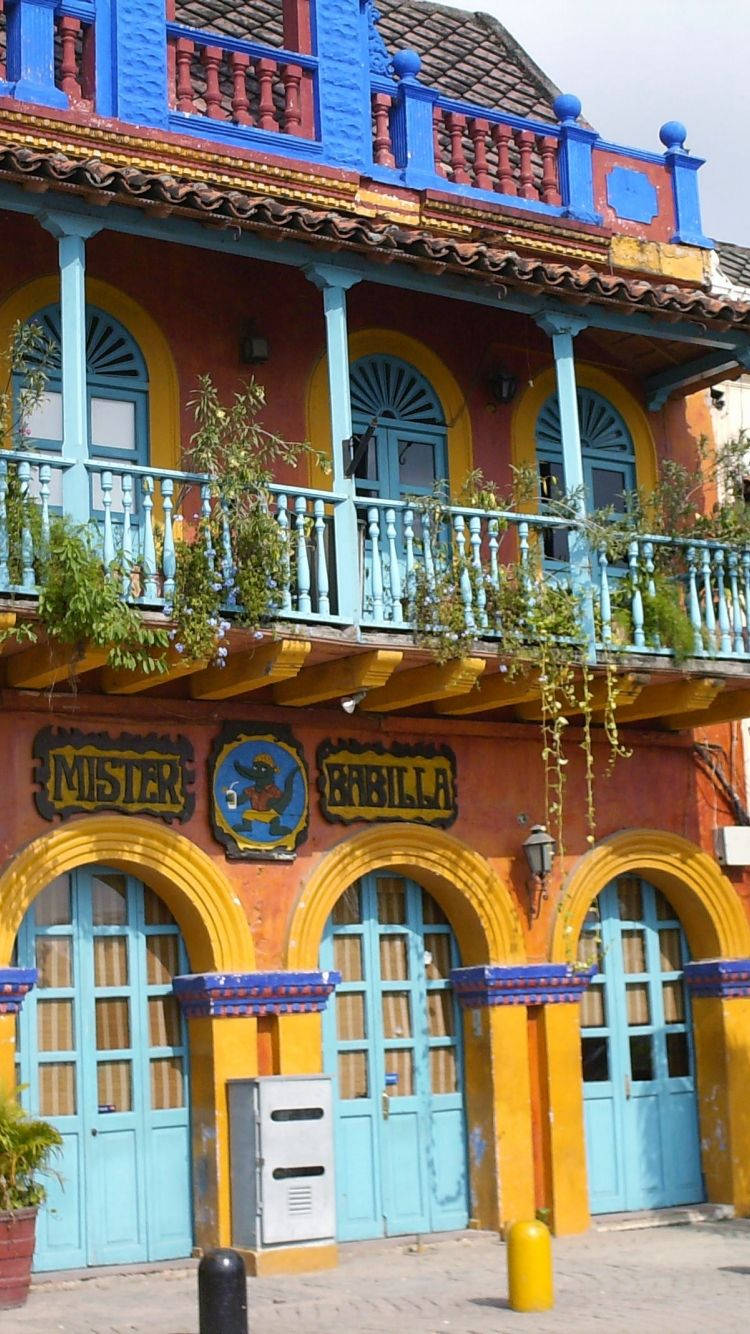 Colombia 桌布 壁紙 — 充滿活力的殖民時期建築，藍色陽台、黃色拱門和窗戶傾瀉而下的綠植，在明亮的日光下格外醒目。