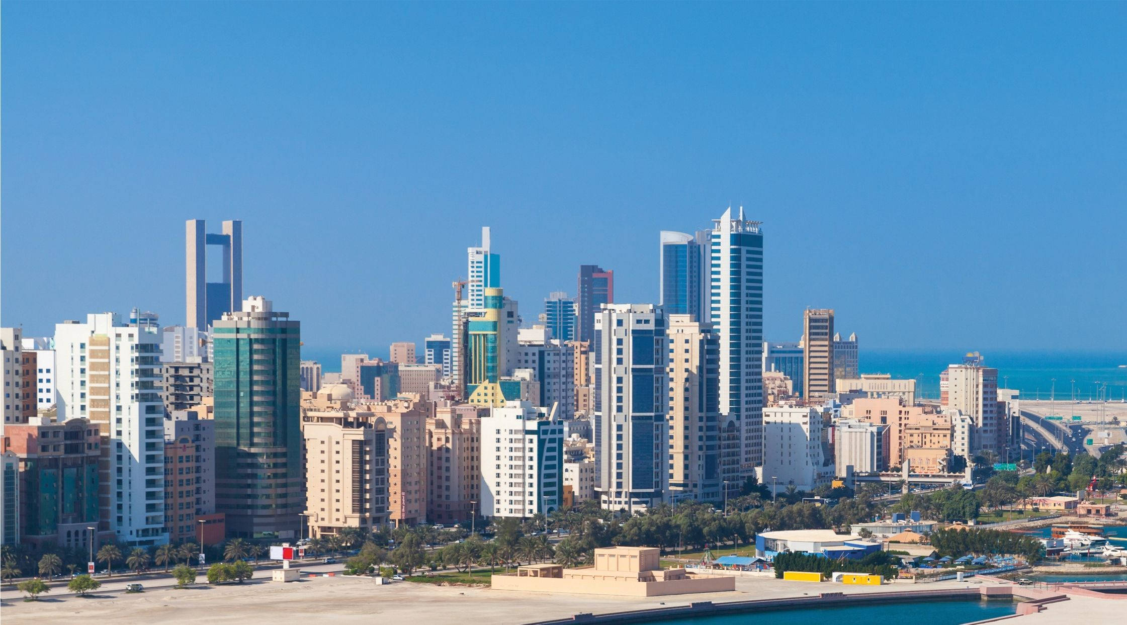 Bahrain 桌布 壁紙 — 充滿活力的沿海城市景觀，現代摩天大樓和低層建築在清澈藍天和海洋背景下矗立。