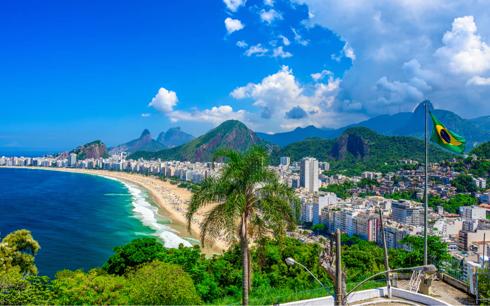 Brazilian Beach 桌布 壁紙 — 充滿活力的沿海城市景觀，綠松石色的海浪拍打著沙灘，沙灘兩旁種滿棕櫚樹和高聳的山脈，在明亮的藍天和白雲下。