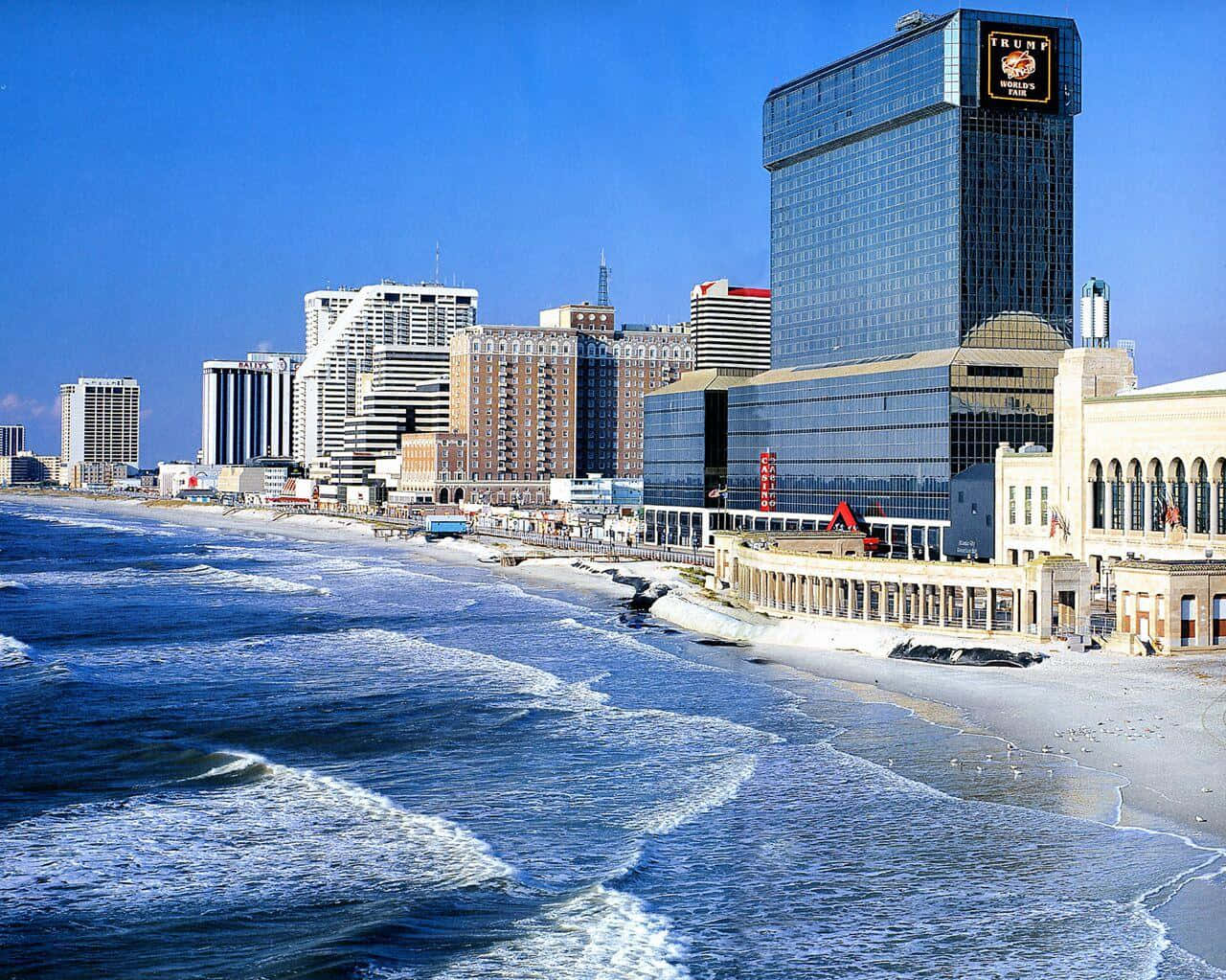 海洋 桌布 壁紙 — 充滿活力的沿海城市景觀，高聳的飯店和賭場沿著沙灘排列，海浪在清澈的藍天下拍打。