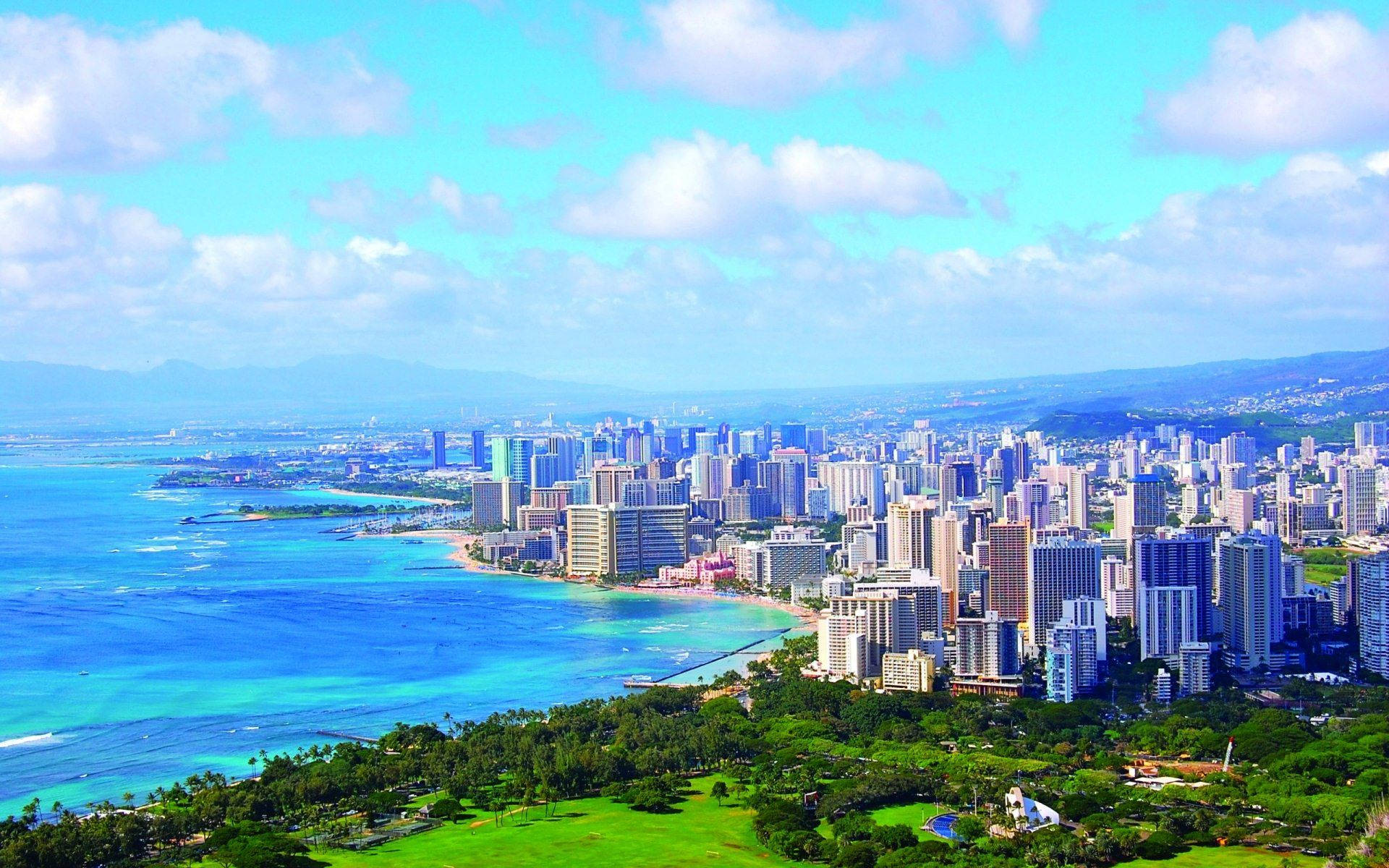 夏威夷 桌布 壁紙 — 充滿活力的沿海城市的鳥瞰圖，擁有碧綠的海洋、鬱鬱蔥蔥的綠色山丘和現代摩天大樓，在明亮的藍天白雲下。