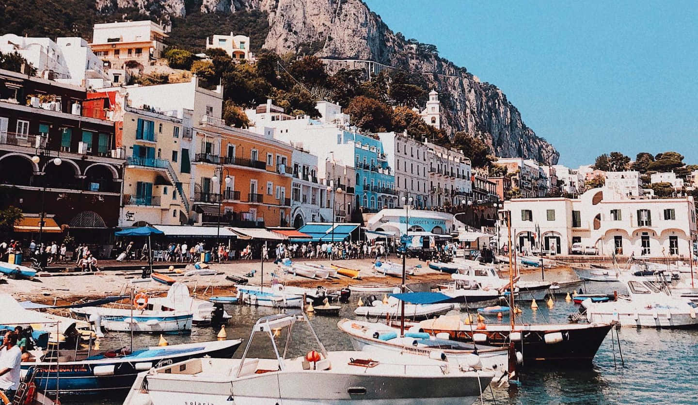 Italian Island 桌布 壁紙 — 充滿活力的沿海小鎮，粉彩建築依附在懸崖上，俯瞰著繁忙的港口，港口停滿船隻，天空清澈湛藍。