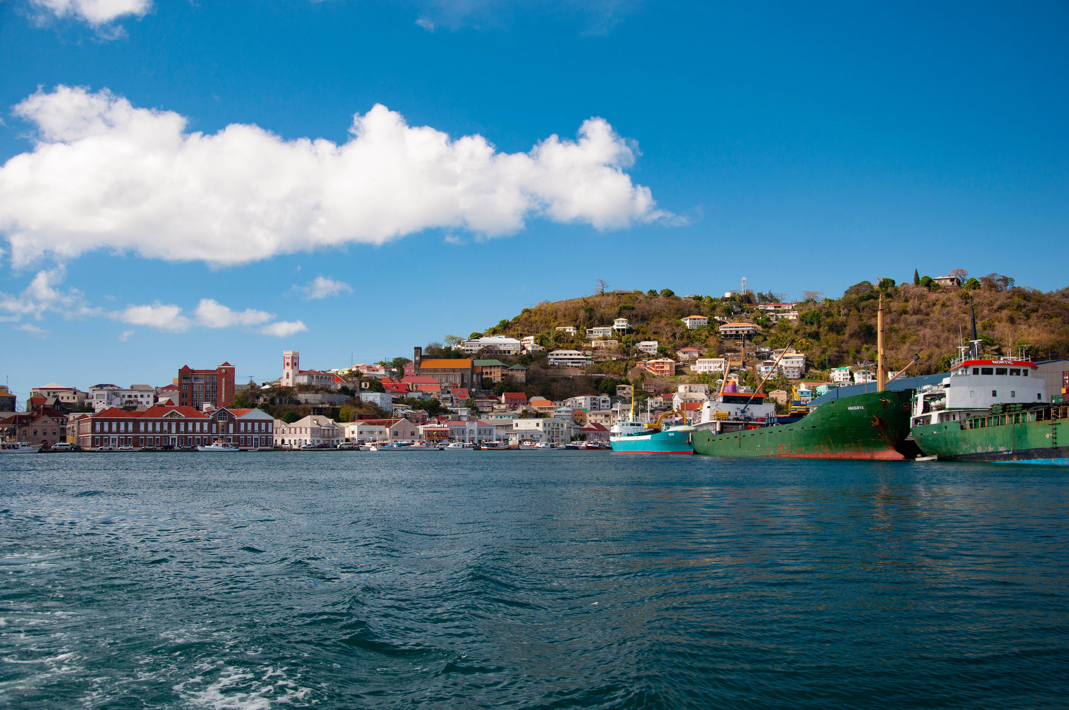 Grenada 桌布 壁紙 — 充滿活力的沿海小鎮，色彩繽紛的建築物依附在山坡上，俯瞰著停滿船隻的港口，上方是明亮的藍天和蓬鬆的白雲。