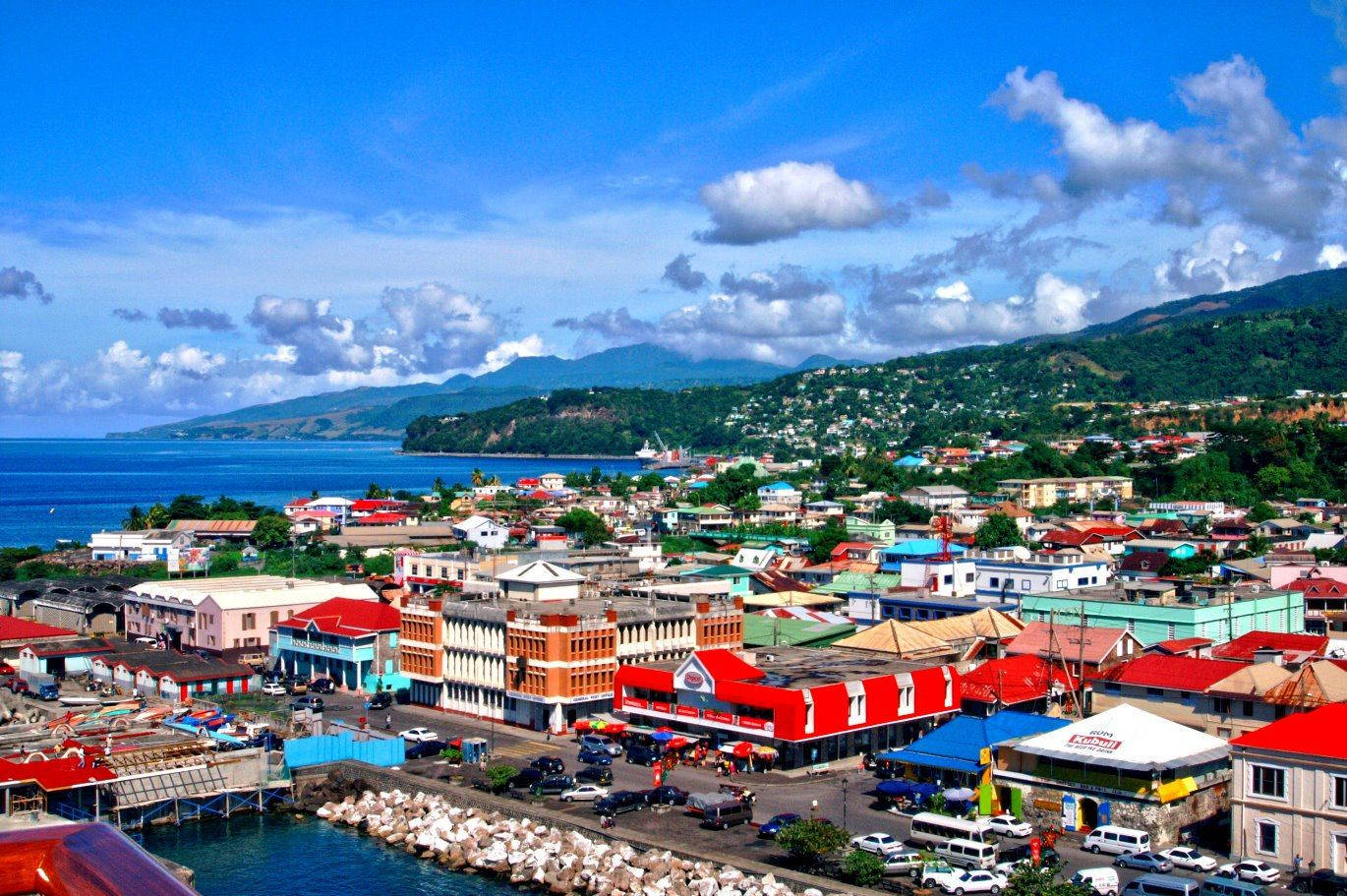 Dominica 桌布 壁紙 — 充滿活力的沿海小鎮，色彩繽紛的建築物坐落在碧綠的海水和鬱鬱蔥蔥的綠色山丘之間，上方是明亮的藍天。