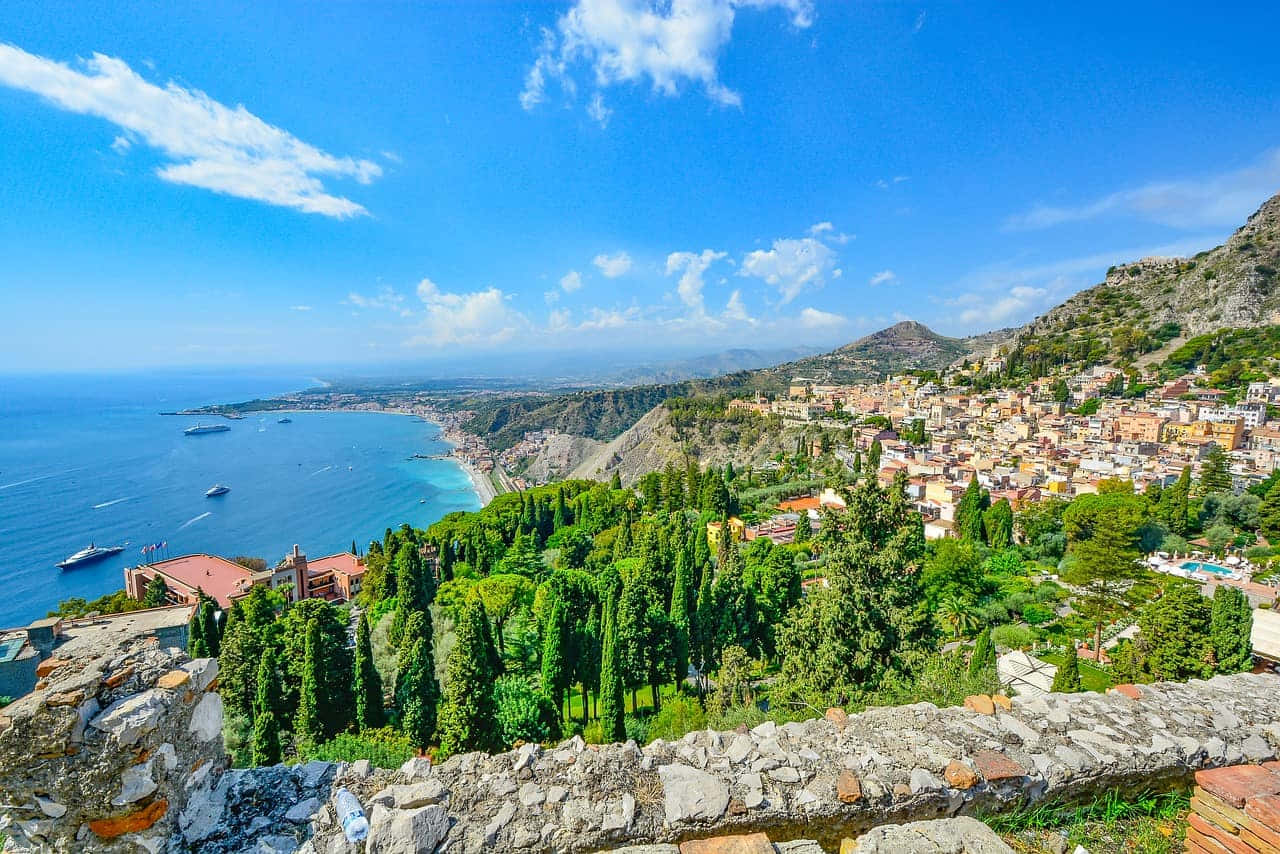 Italian Island 桌布 壁紙 — 充滿活力的沿海小鎮依附在陽光普照的懸崖上，俯瞰著蔚藍的天空下散落著雲朵的碧綠大海。