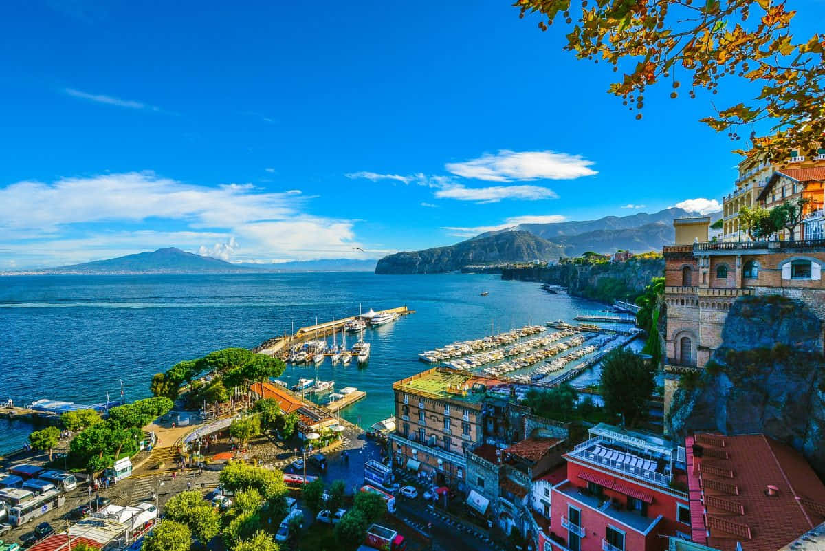 Italian Island 桌布 壁紙 — 充滿活力的沿海小鎮緊貼懸崖，俯瞰著閃閃發光的藍色海灣，海灣內有碼頭和遠處的山脈，天空晴朗。