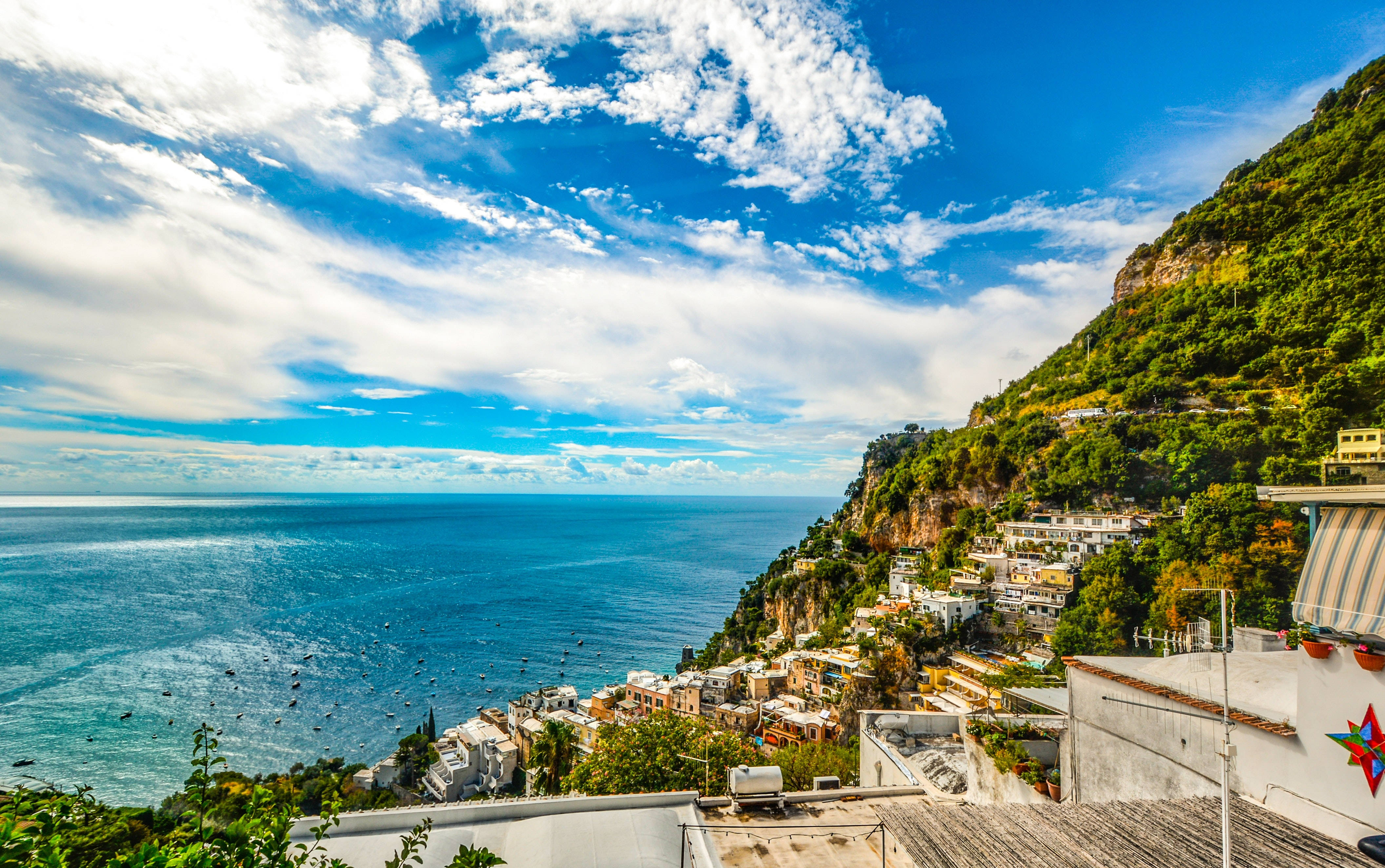 Amalfi Coast 桌布 壁紙 — 充滿活力的沿海村莊緊貼在陡峭的綠色懸崖上，俯瞰廣闊的藍色大海，天空明亮，雲朵蓬鬆。