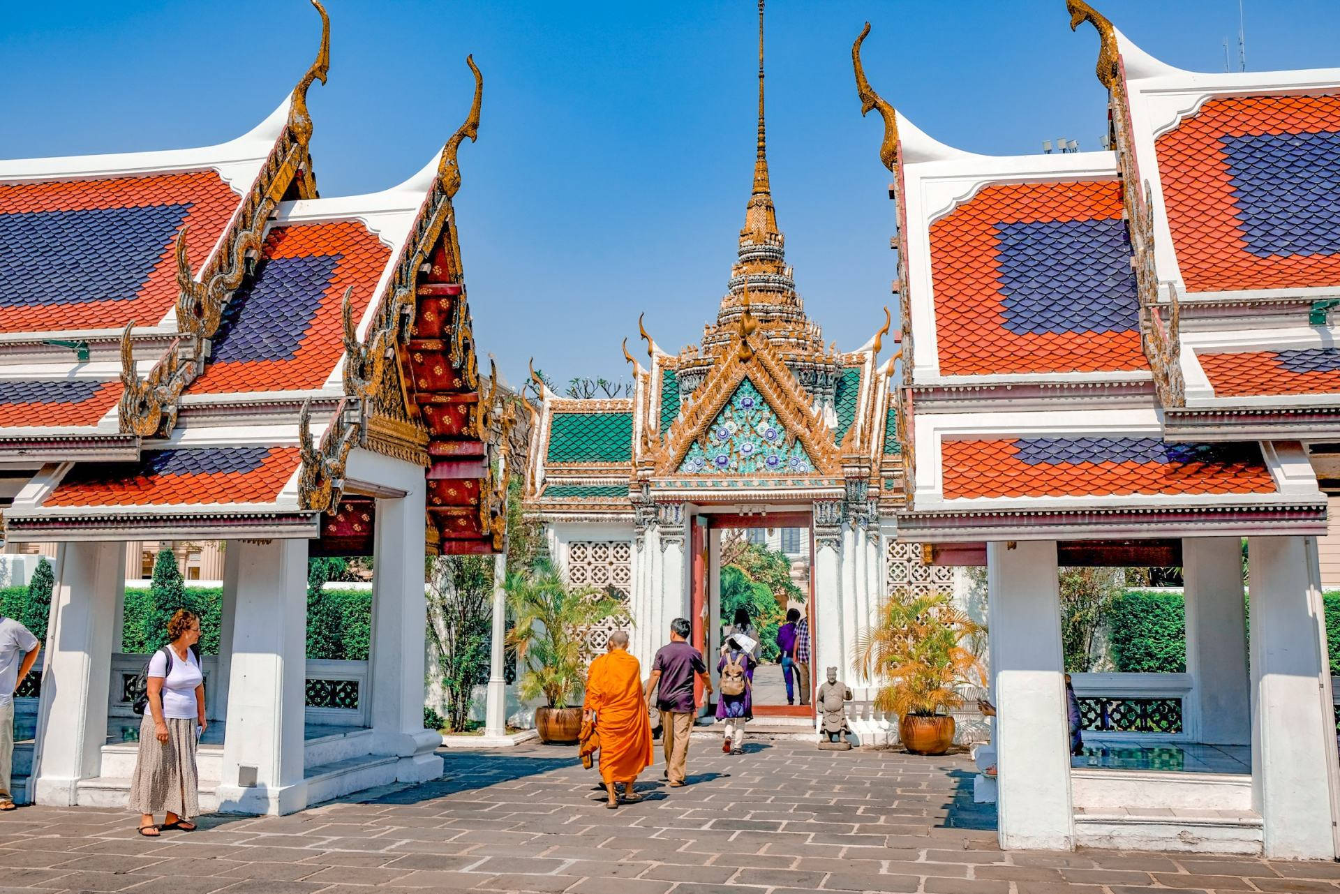 Bangkok 桌布 壁紙 — 充滿活力的泰國寺廟庭院，裝飾華麗的紅色和金色屋頂，在晴朗的藍天下，遊客穿過入口。
