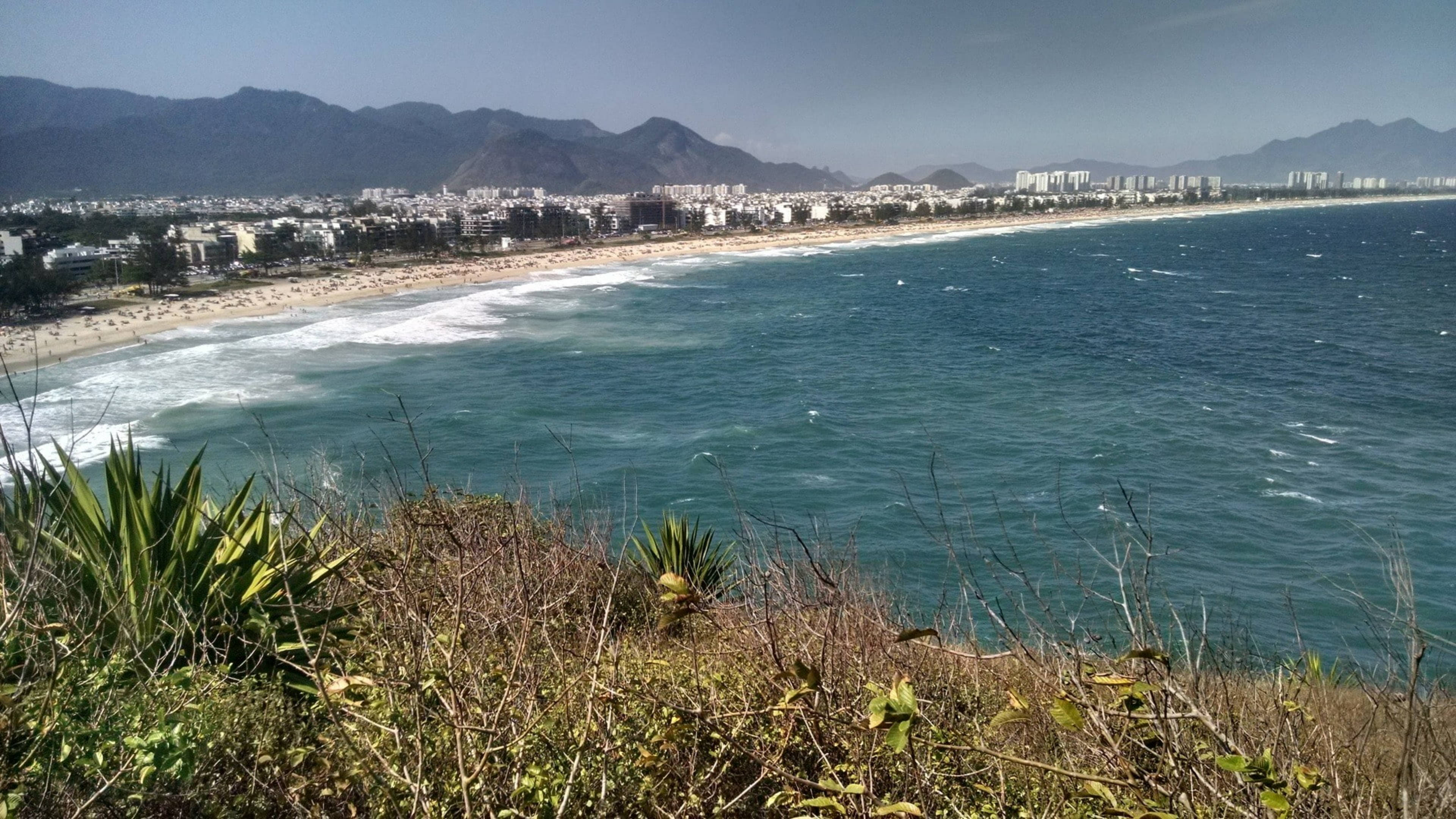 rio 桌布 壁紙 — 充滿活力的海岸景色，在晴朗的天空下，綠松石色的海浪拍打著沙灘，沙灘兩旁是建築物和遠處的山脈。