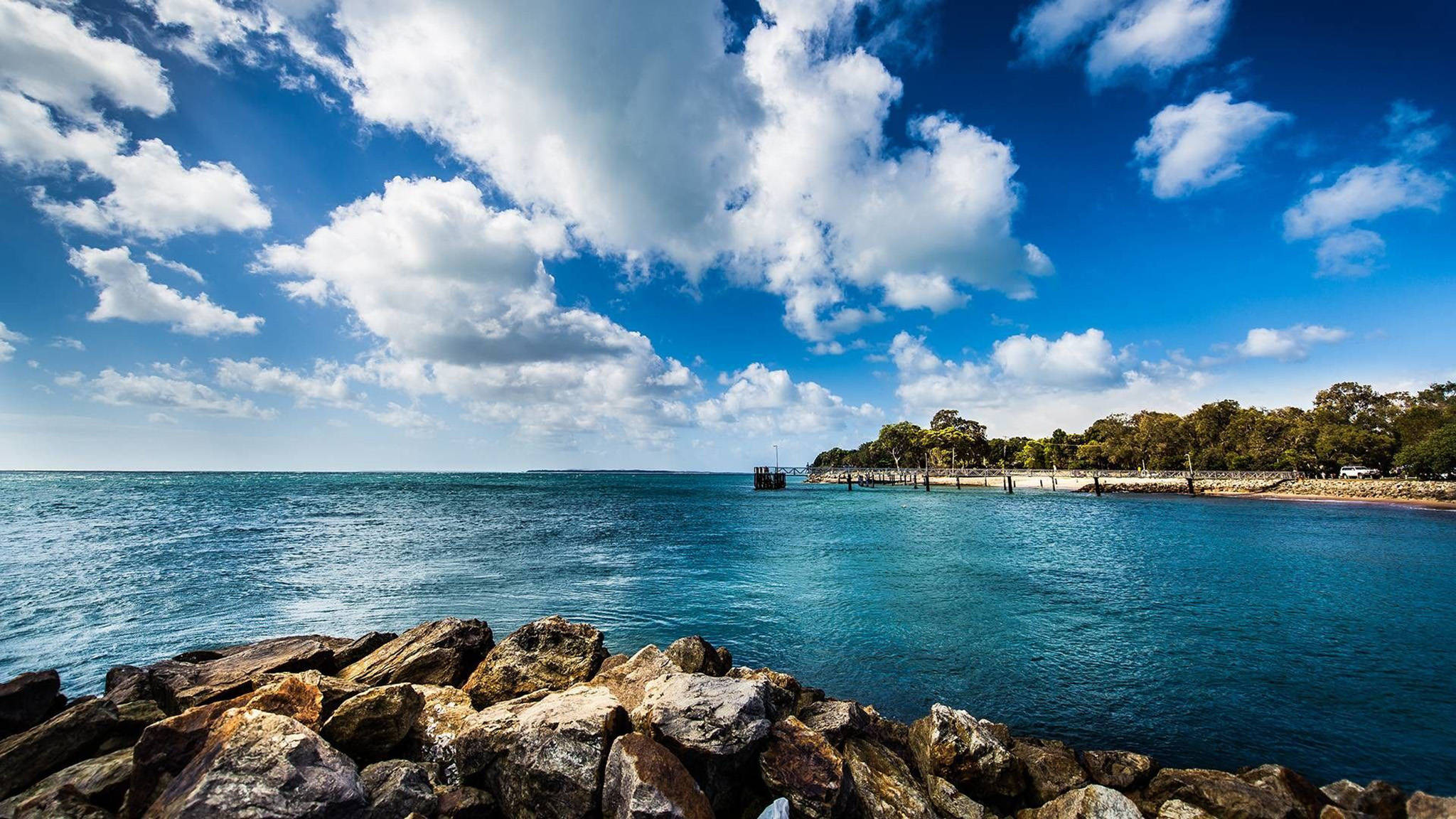 View 桌布 壁紙 — 充滿活力的海岸景色，有碧綠的海水、岩石前景和茂密的綠樹，在明亮的藍天下有蓬鬆的白雲。