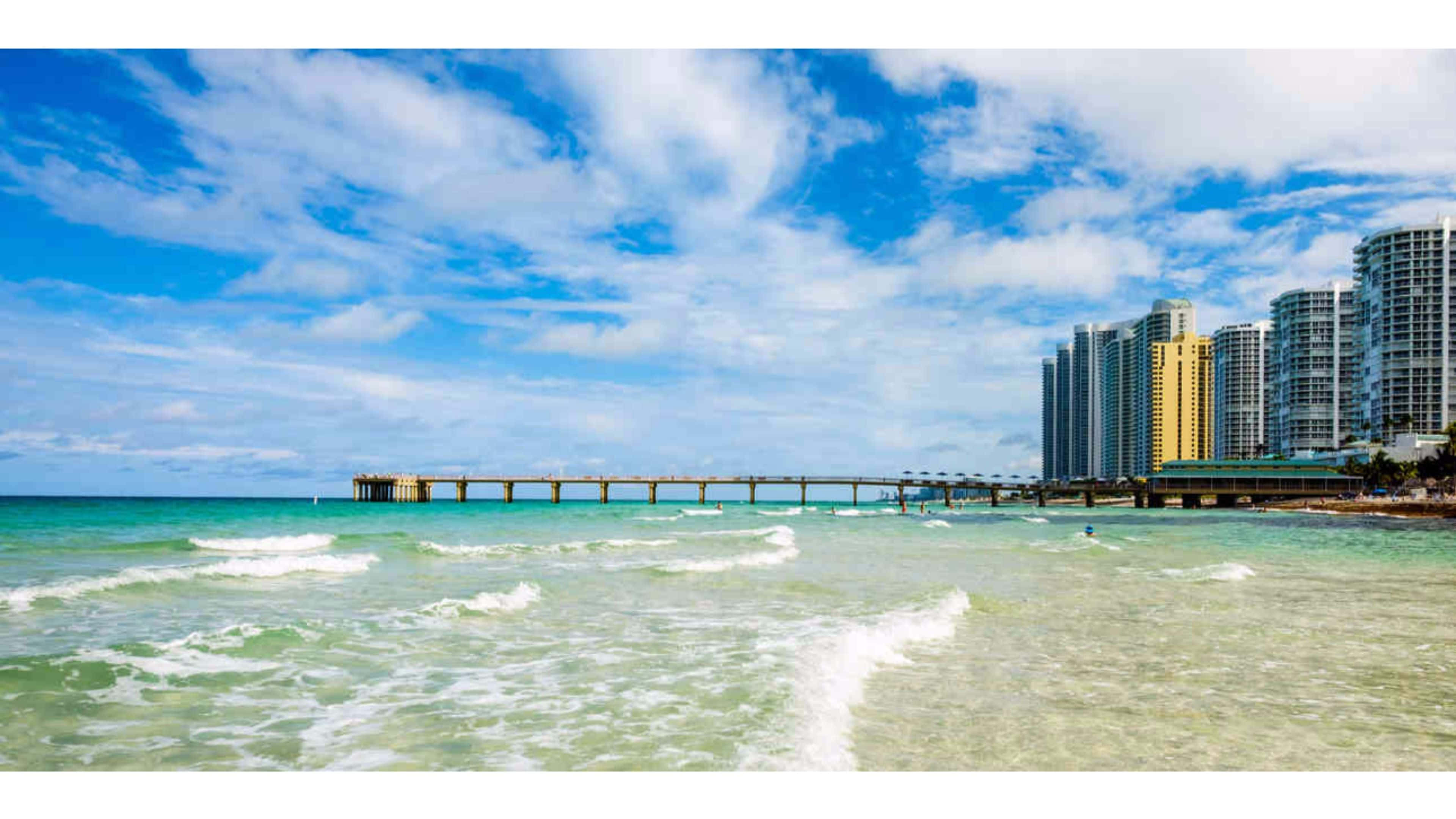 Miami 4K 桌布 壁紙 — 充滿活力的海岸景色，綠松石色的海浪在木製碼頭附近拍打，背景是現代高層建築，在明亮的藍天和蓬鬆的白雲下。