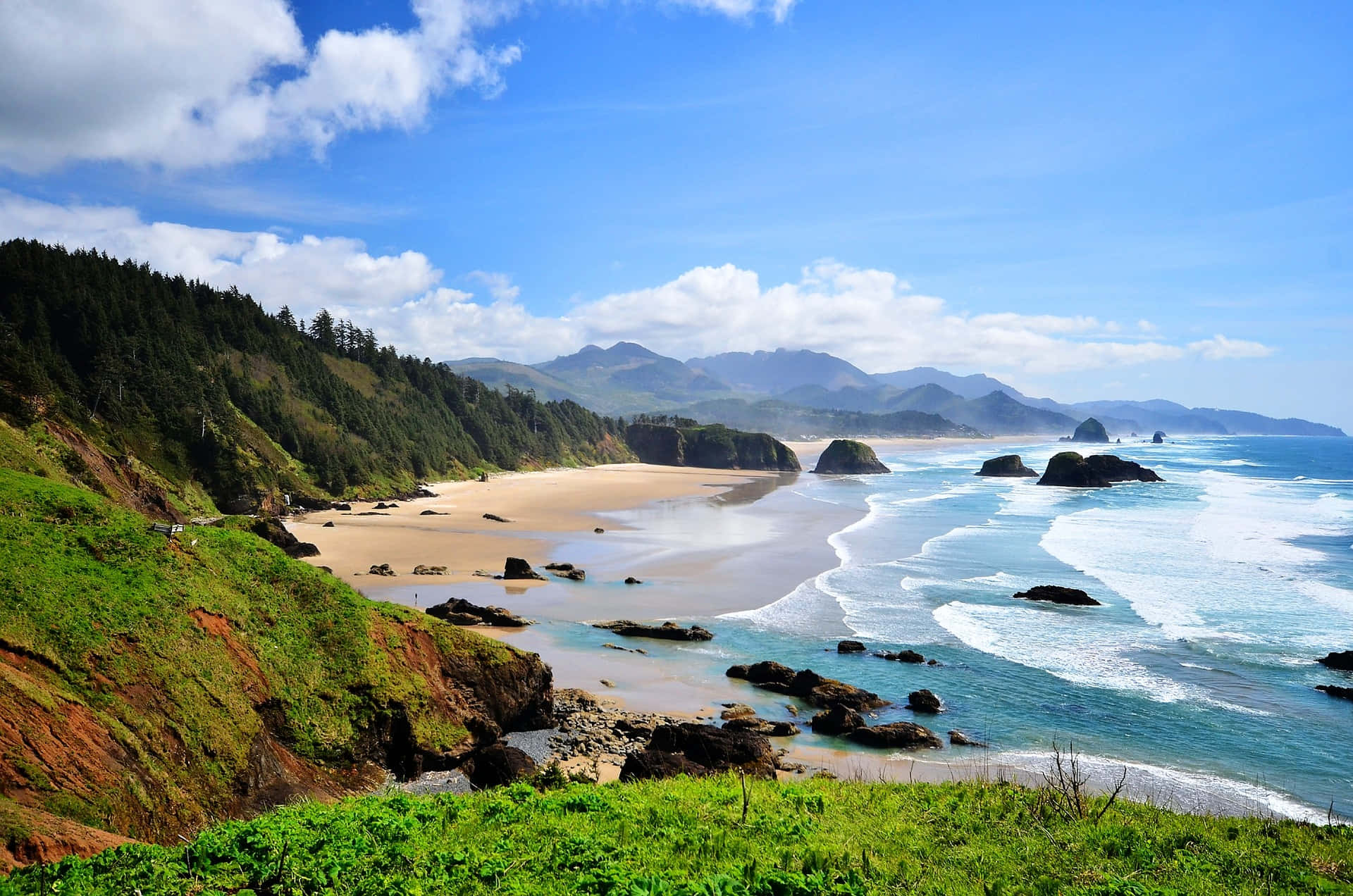 Oregon Coast 桌布 壁紙 — 充滿活力的海岸景色，綠松石色的海浪拍打著沙灘，四周環繞著茂密的綠色懸崖和遠處的山脈，在明亮的藍天下。