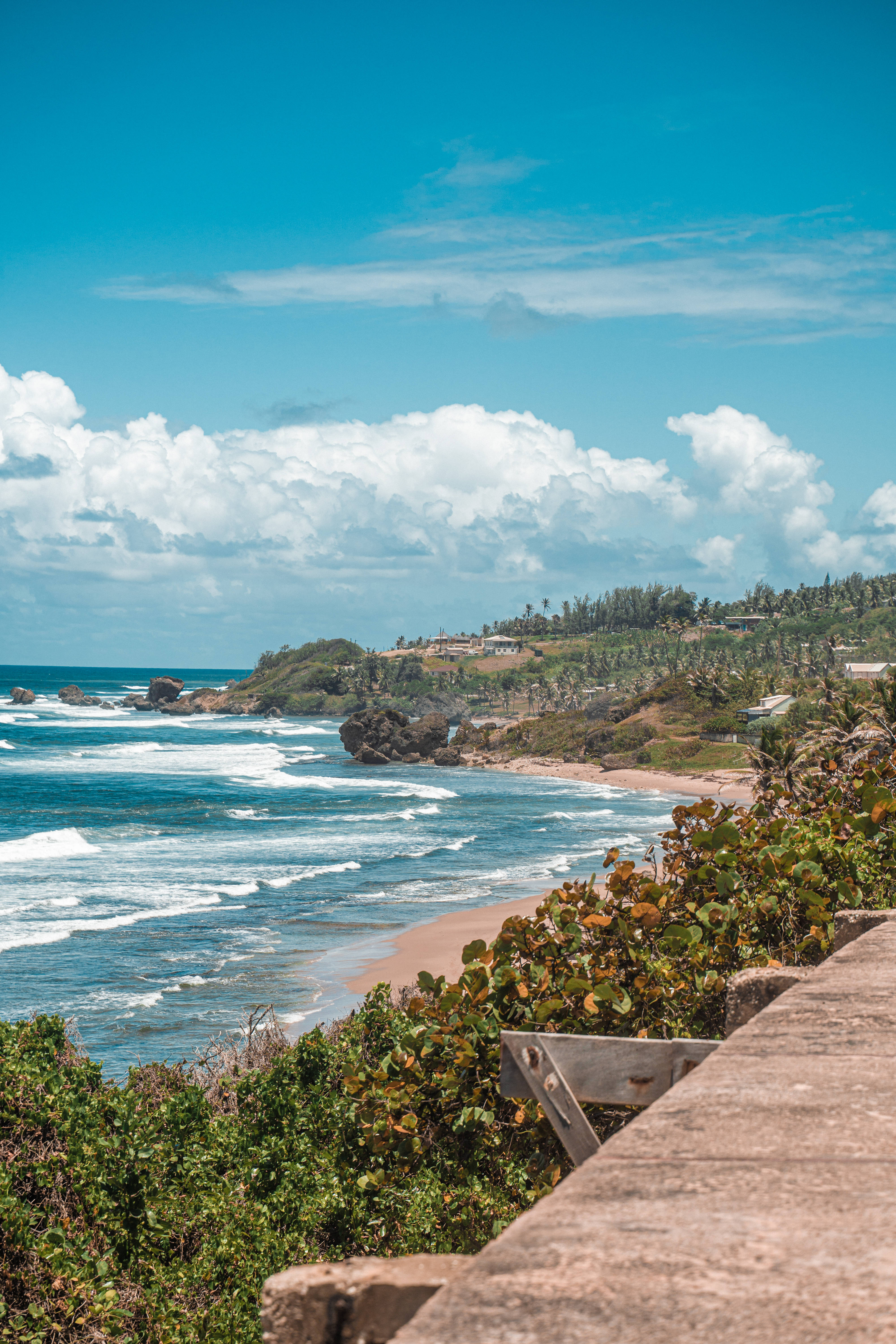 Barbados 桌布 壁紙 — 充滿活力的海岸景色，綠松石色的海浪拍打著沙灘，四周環繞著茂密的綠色山丘和木製欄杆，上方是明亮的藍天和蓬鬆的白雲。
