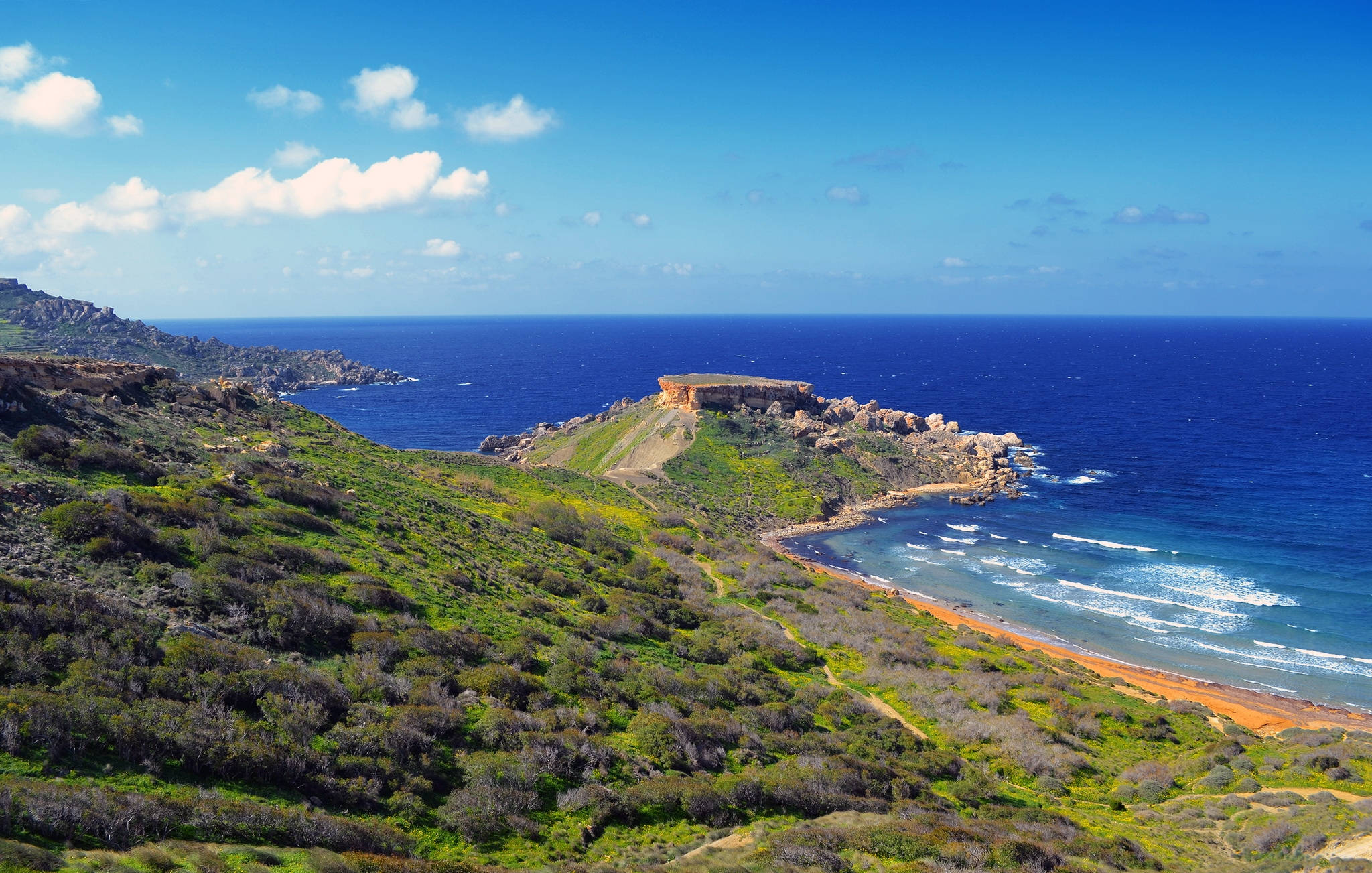 Malta 桌布 壁紙 — 充滿活力的海岸景觀，鬱鬱蔥蔥的綠色山丘與碧綠的海水相遇，在明亮的藍天下有散落的雲朵。
