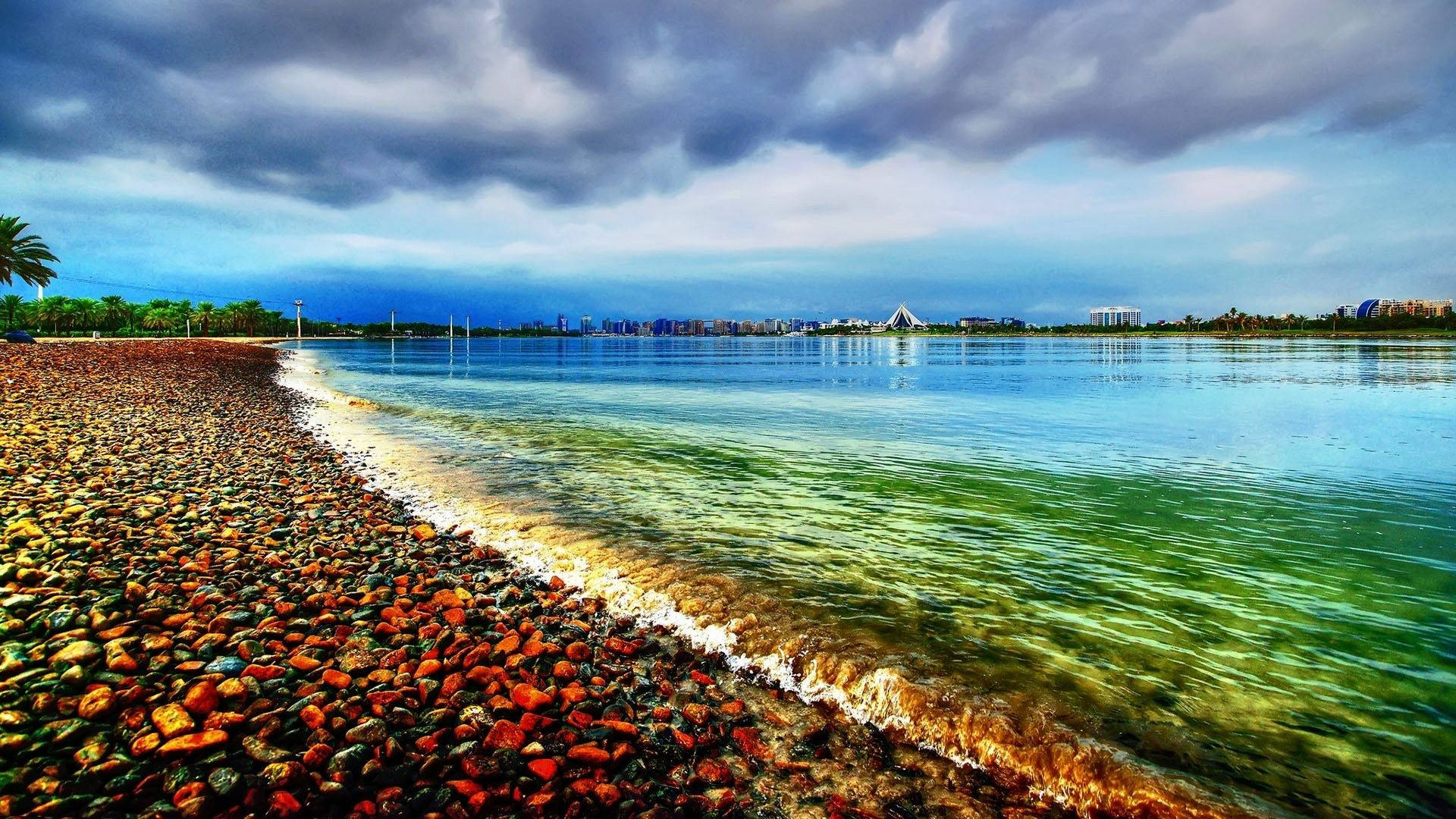 海灘 桌布 壁紙 — 充滿活力的海岸線，色彩繽紛的鵝卵石與碧綠的海水相遇，在戲劇性的多雲天空下，遠處有城市景觀。