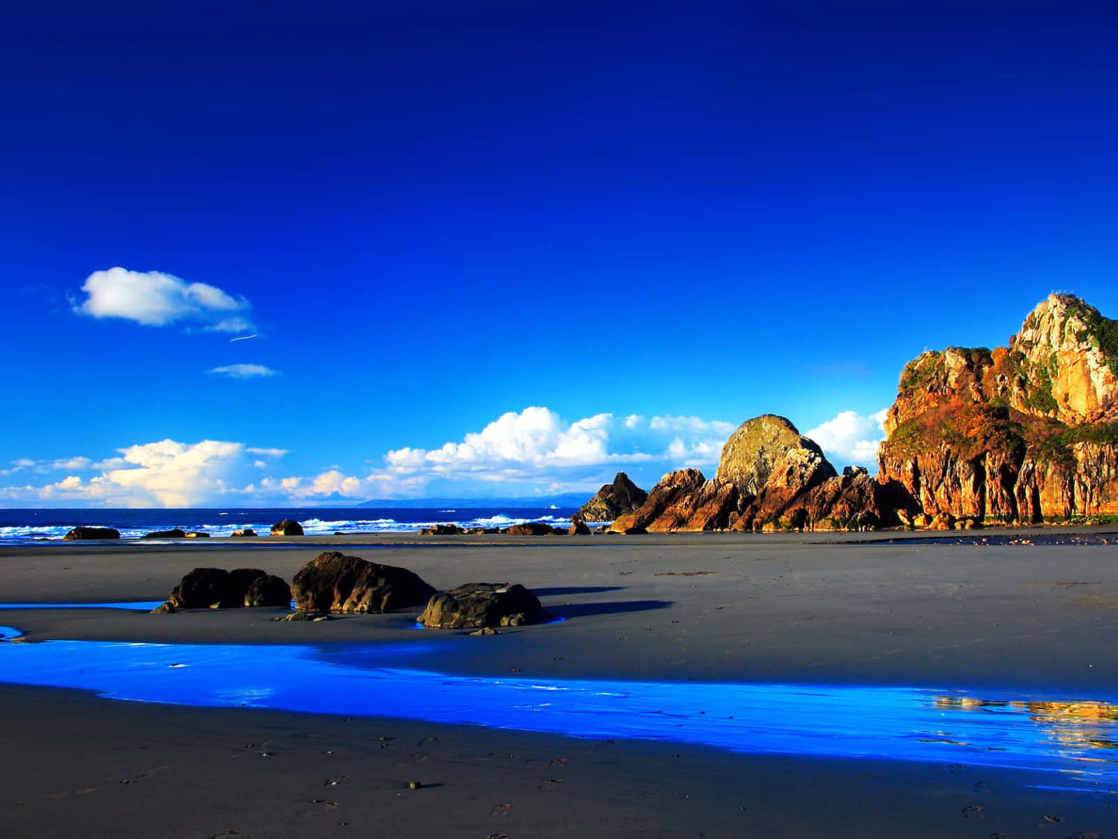 Blue Sky 桌布 壁紙 — 充滿活力的海灘場景，有深色沙灘、發光的藍色潮汐池和崎嶇的橙色懸崖，在深藍色的天空下，有蓬鬆的白雲。